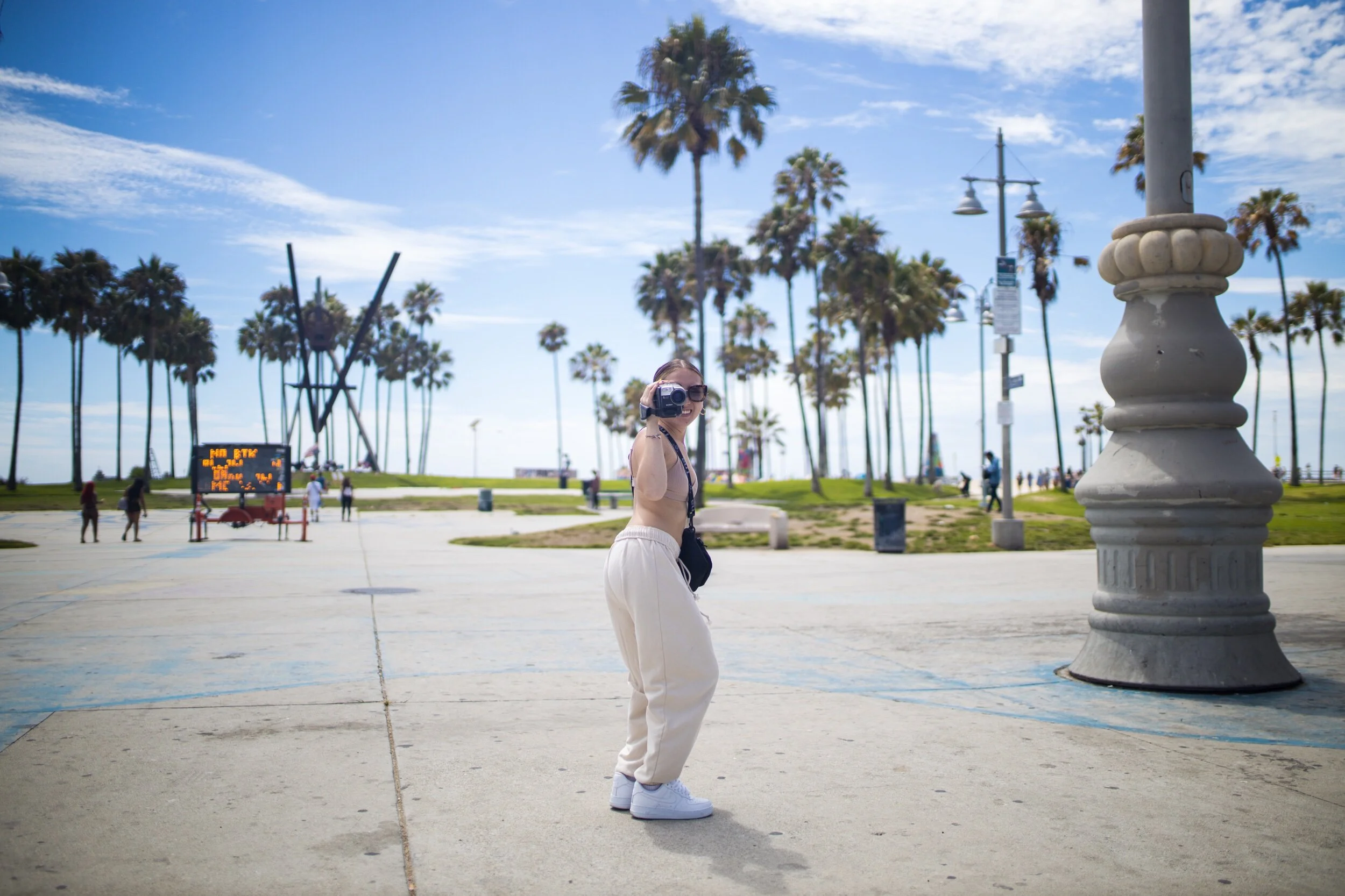 A woman in beige pants and white sneakers is standing on a sunny boardwalk near palm trees, holding a vintage camera, with a bright blue sky overhead.