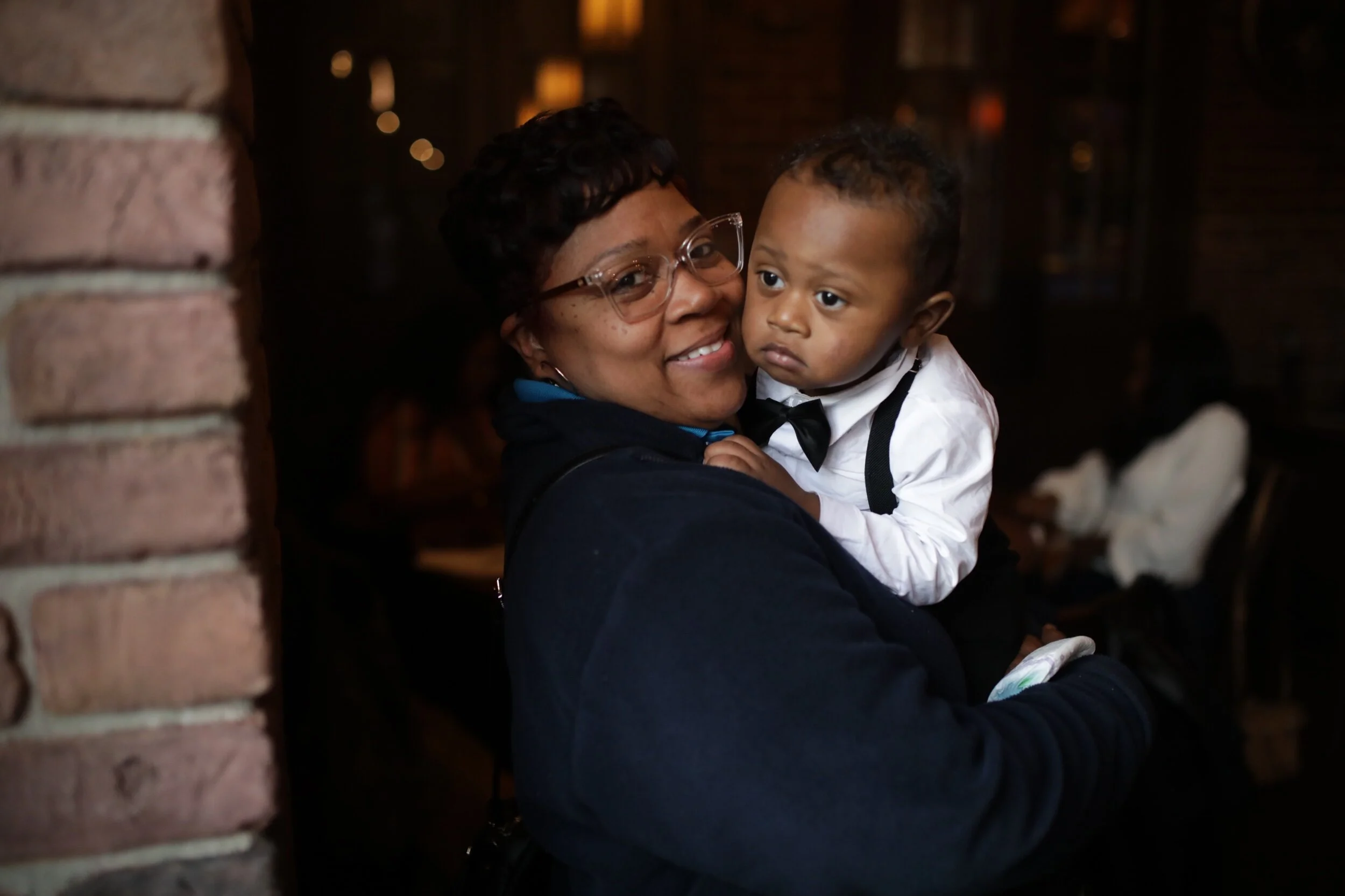 A woman holding a young boy dressed in a white shirt and black bow tie, smiling, in a warmly lit indoor setting.