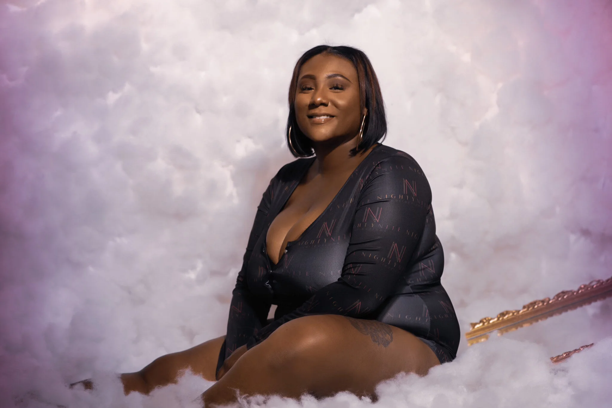 A woman with shoulder-length dark hair, hoop earrings, and a black patterned dress, sitting among white clouds with a pastel pink and purple background.