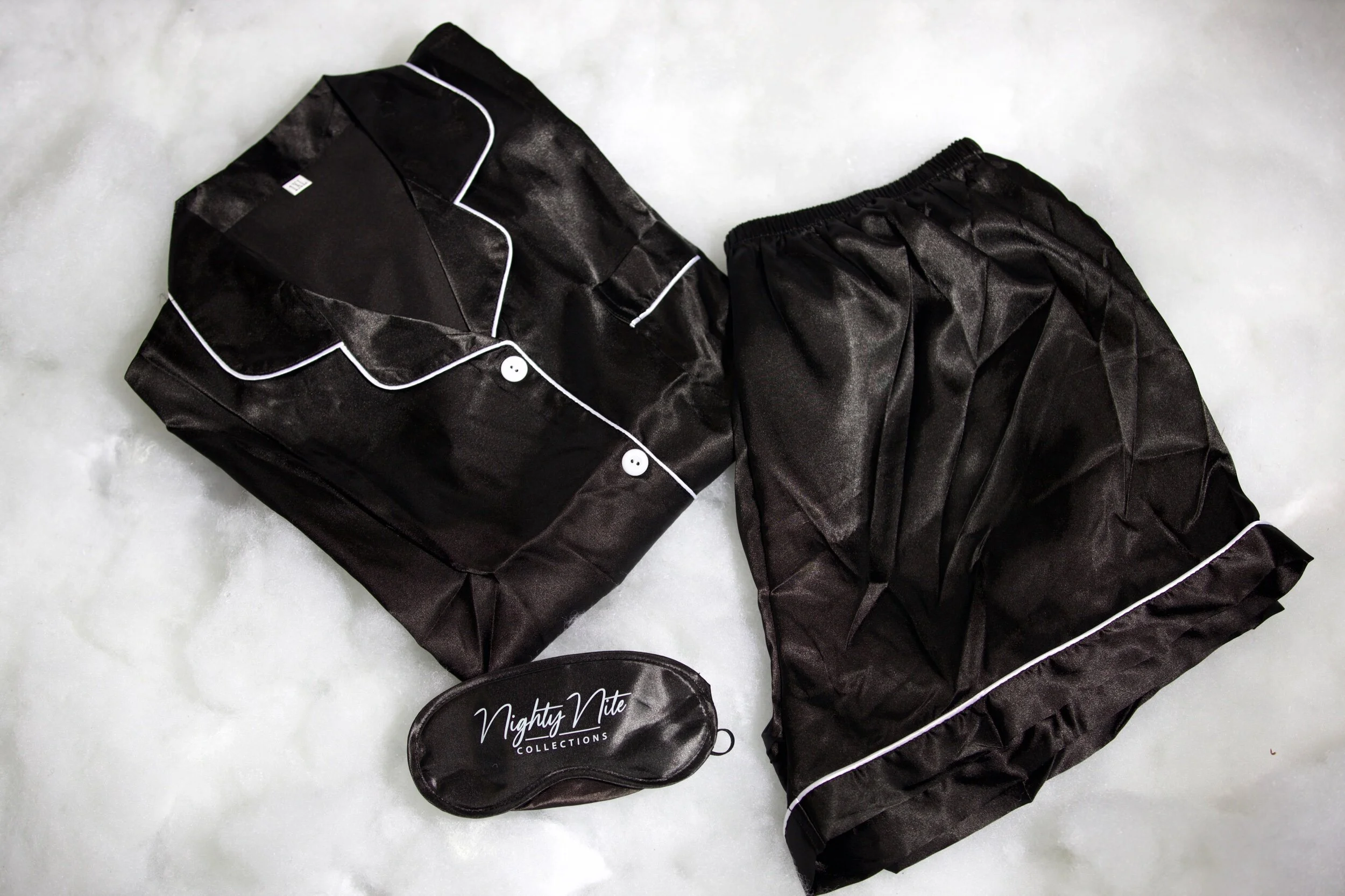 Black satin pajamas with white piping, folded on a light surface, with a black sleep mask that says 'Nighty Nite Collections'.
