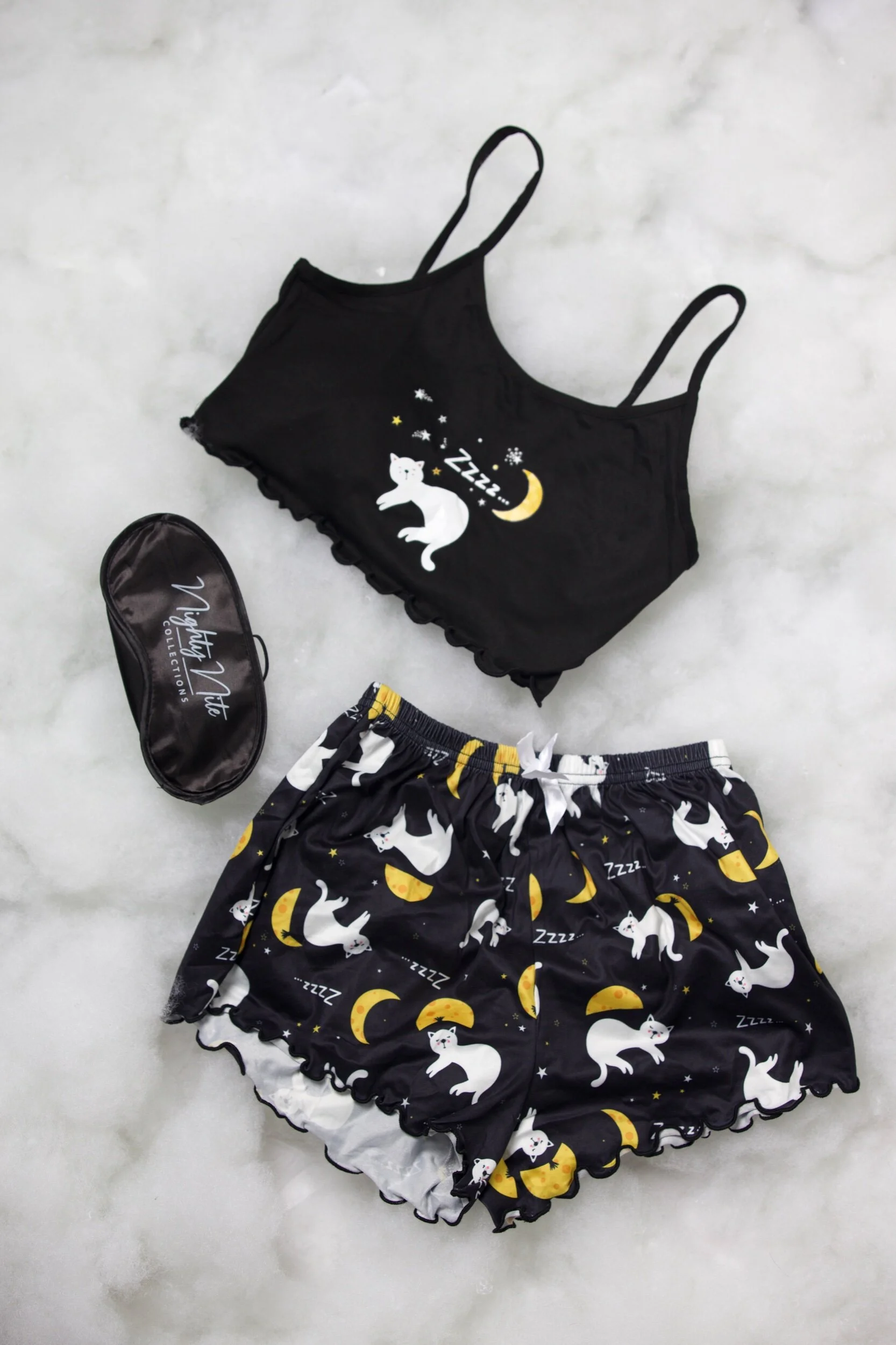 Sleepwear set with a black camisole top and matching shorts featuring a white cat, moon, and stars pattern, along with a black eye mask with matching pattern, on a light gray surface.