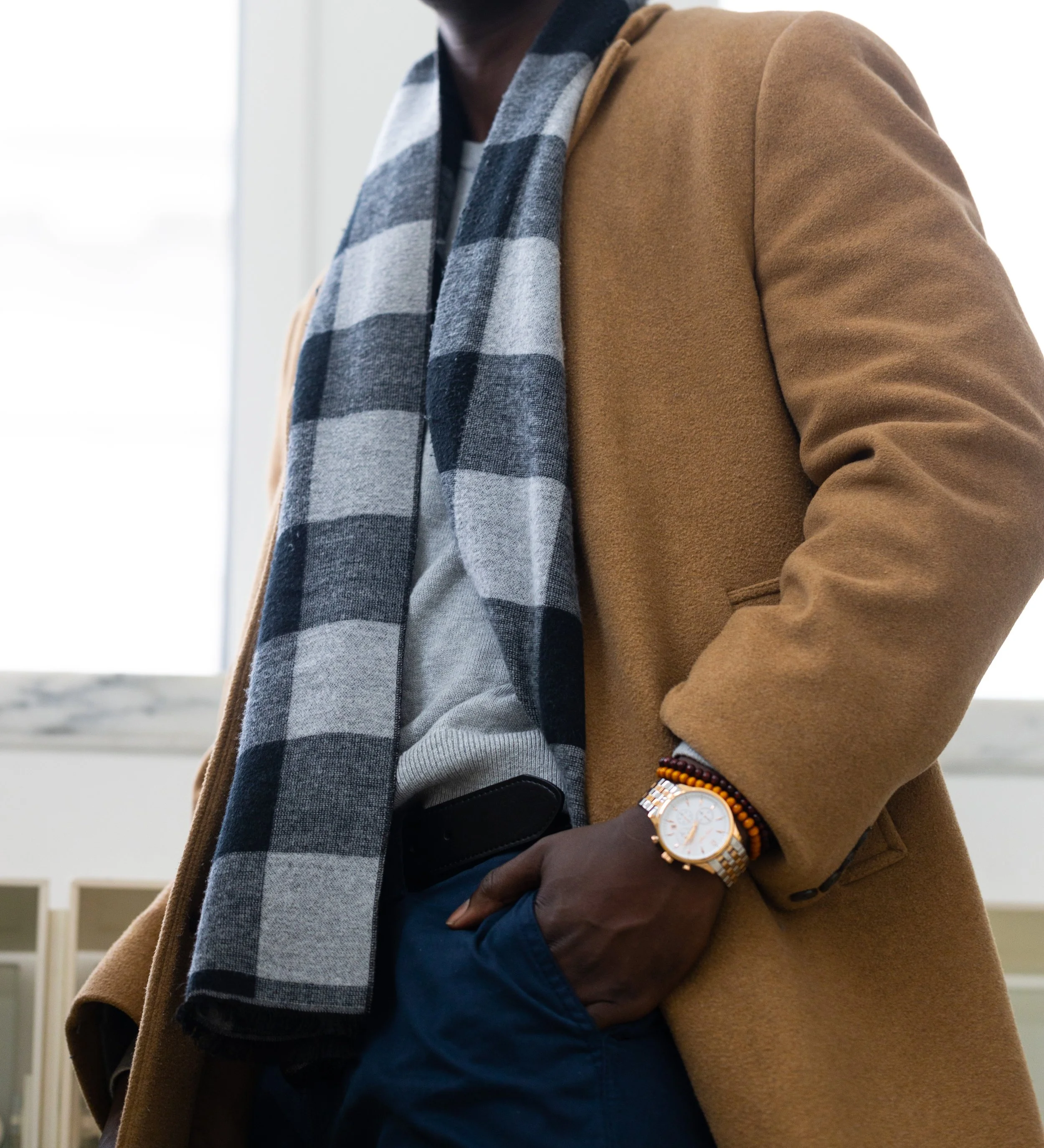 Close-up of a person wearing a camel coat, gray shirt, blue pants, a striped scarf, and a watch with a gold band, standing indoors near a window.