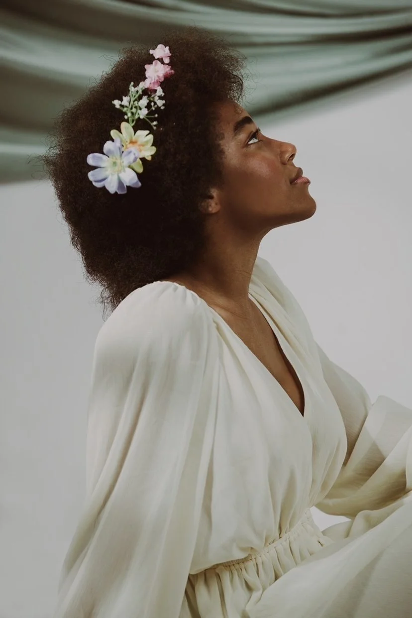 A woman with dark skin and voluminous curly hair styled with a floral headband, wearing a cream-colored dress with puffed sleeves, looking upward with a serene expression.