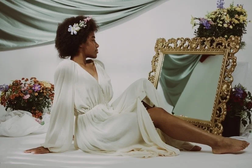 A woman with a flower in her hair, wearing a long white dress, sitting on a bed surrounded by flowers, with a large ornate mirror and floral arrangements in the background.