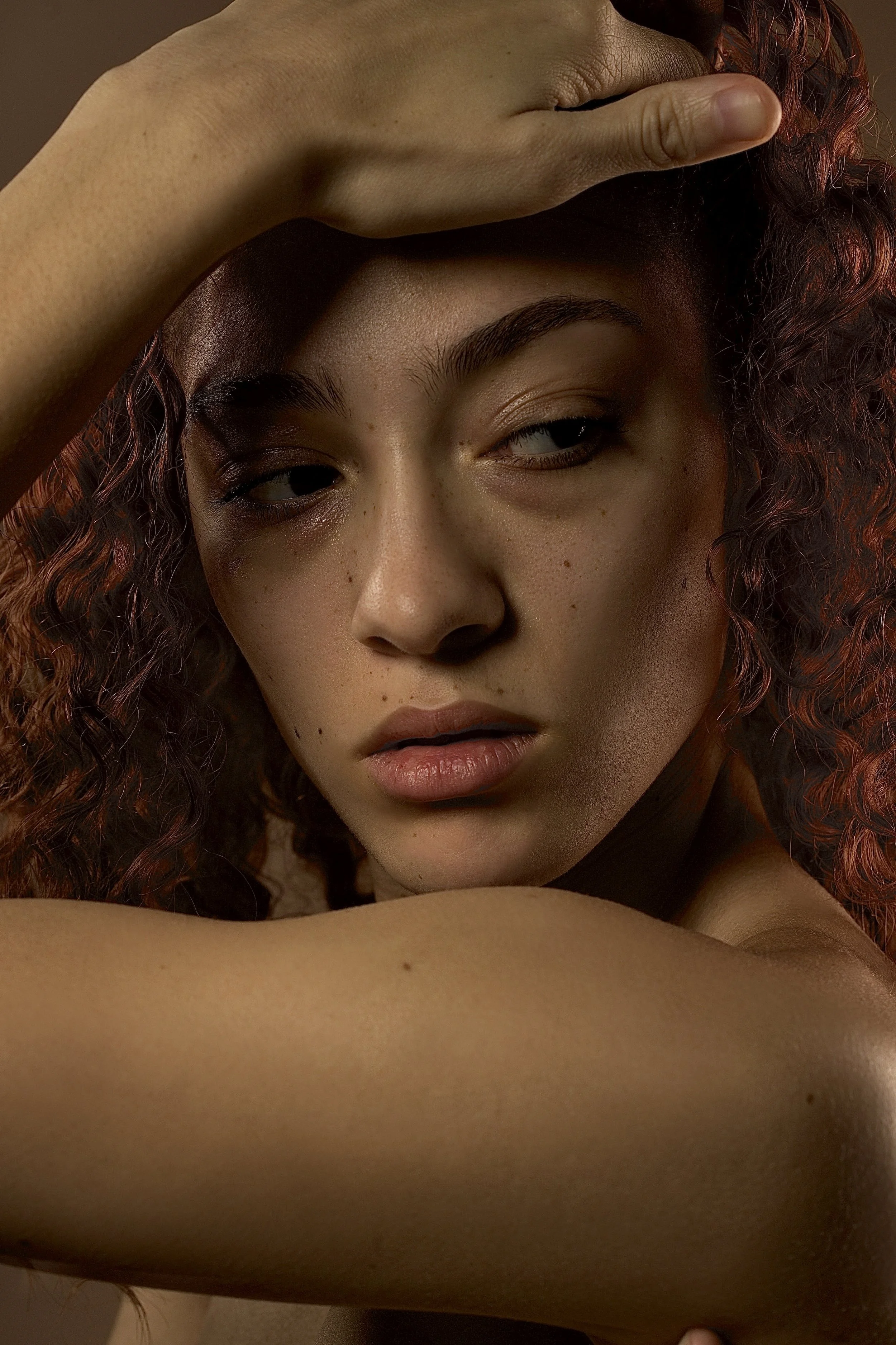 Close-up portrait of a woman with curly red hair, resting her arm on her forehead, with a contemplative expression.
