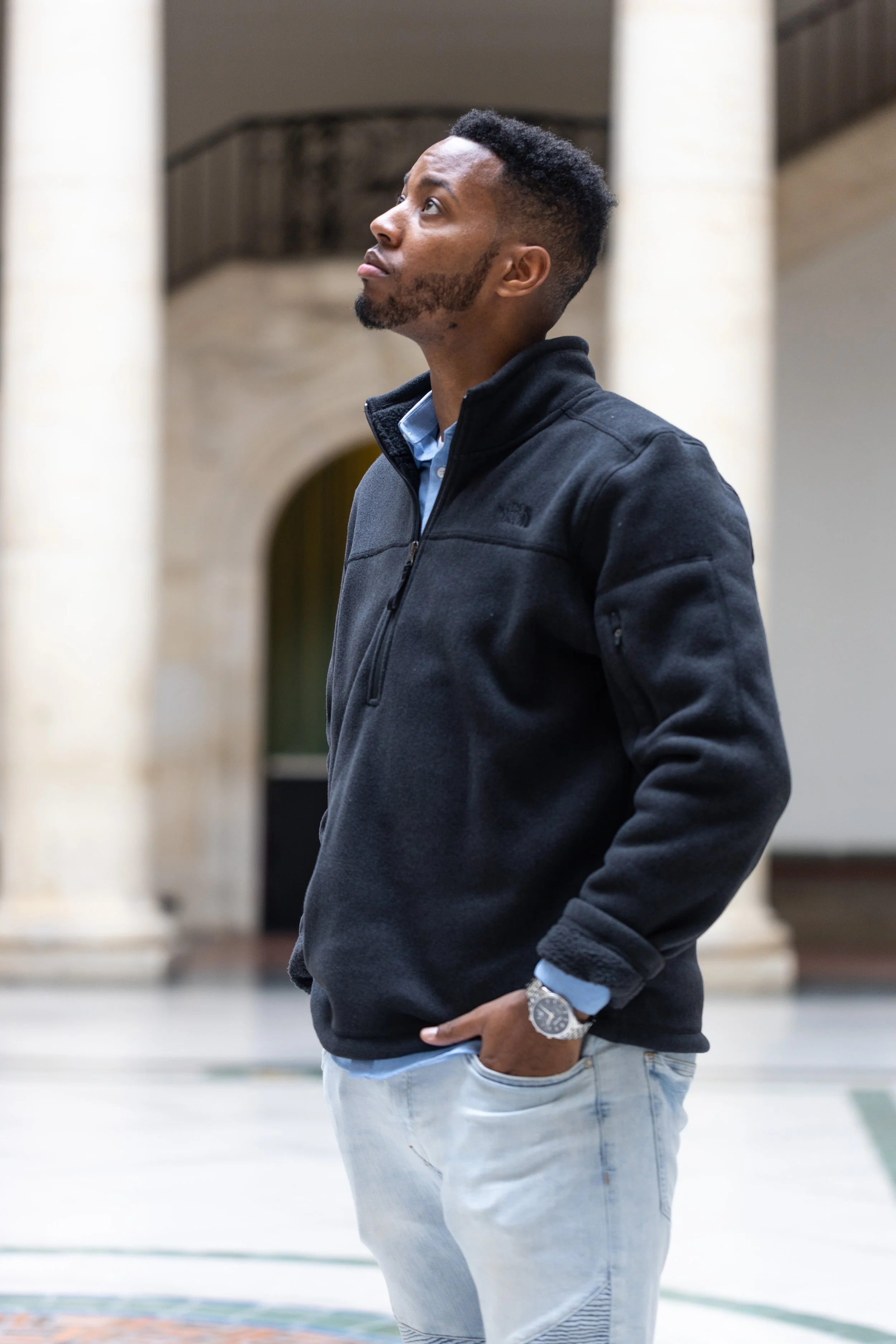 A young man with short hair and a beard looking up, wearing a black North face jacket, light blue shirt, light jeans, and a watch, standing indoors near columns.