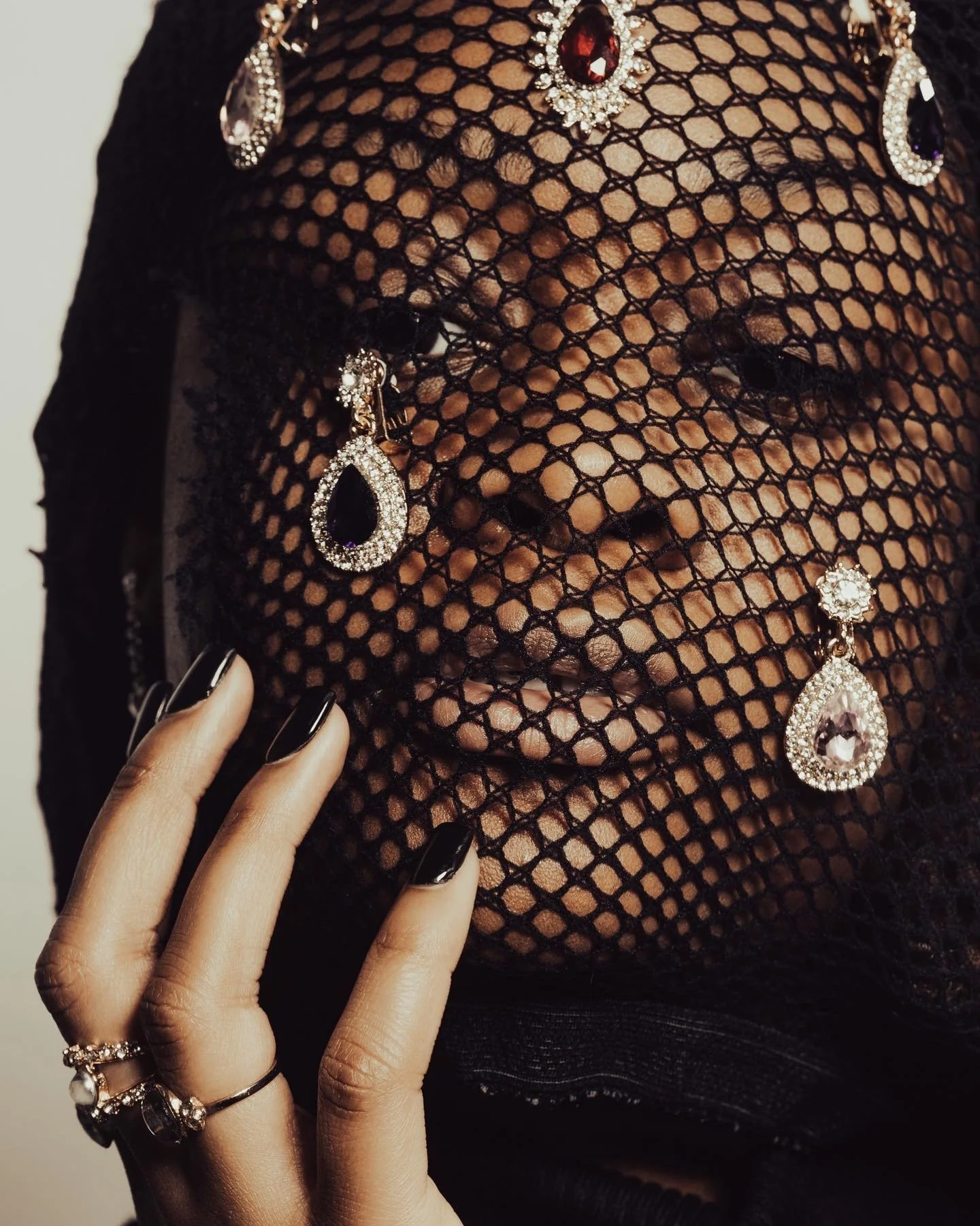 A close-up of a person's face partially covered by a black fishnet fabric, adorned with large, sparkling earrings and a matching necklace with red and purple gemstones. The person’s hand, with dark nail polish and multiple rings, is gently touching t