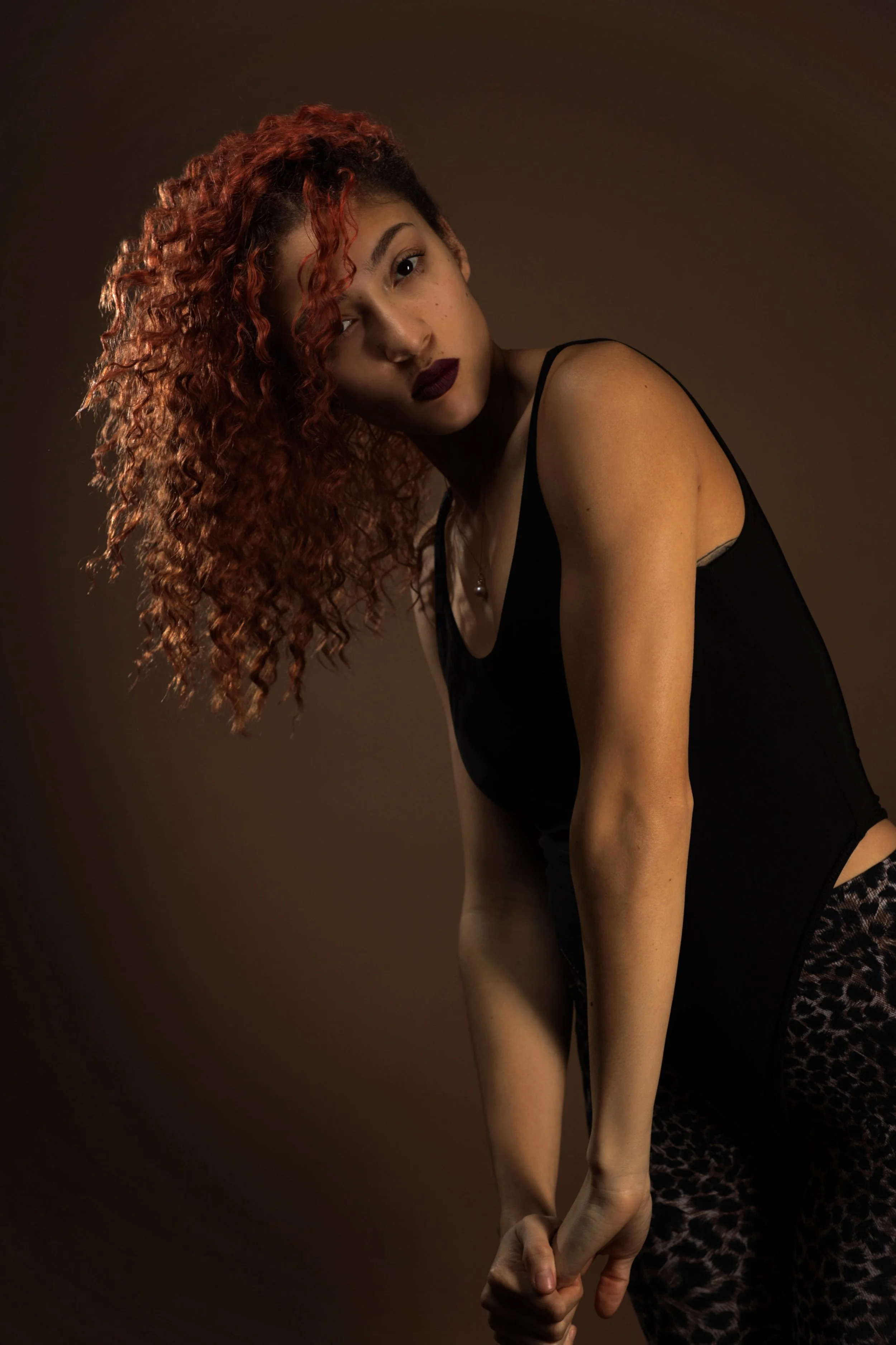 A woman with curly red hair posing against a dark background, wearing a black tank top and leopard-print pants.