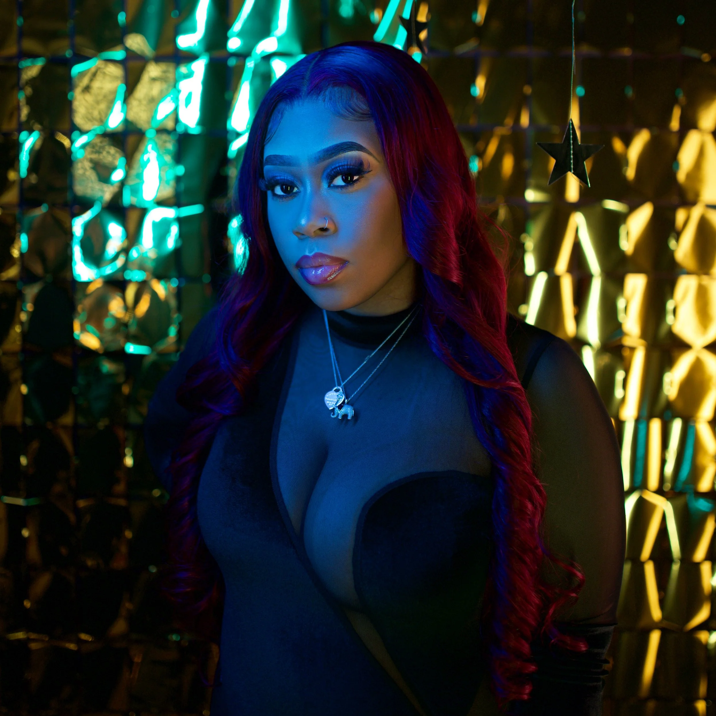 A woman with long wavy hair, colored blue and purple, posing against a background of colorful neon lights and hanging star decorations.