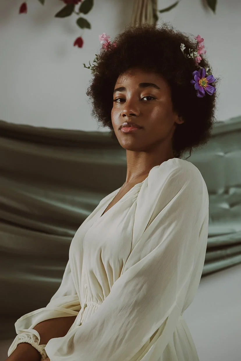 A woman with natural curly hair decorated with pink and purple flowers, wearing a cream-colored blouse, standing in front of a draped fabric backdrop.