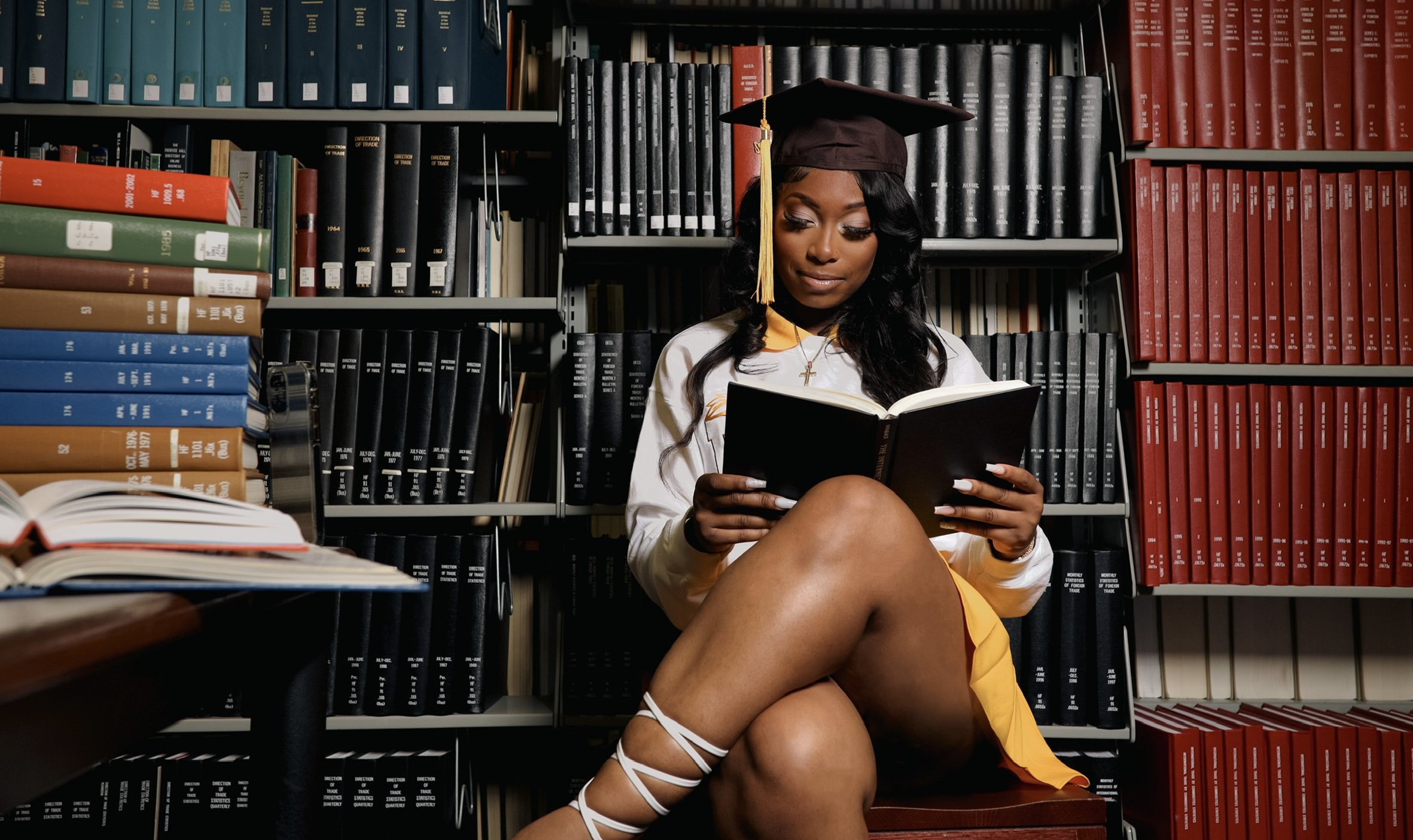A woman in a graduation cap and gown sitting on a bench in a library, reading a book, surrounded by shelves of books.