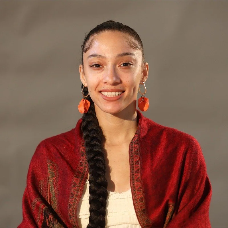 Young woman with long braided dark hair, wearing large orange earrings and a red shawl over a white blouse, smiling at the camera.