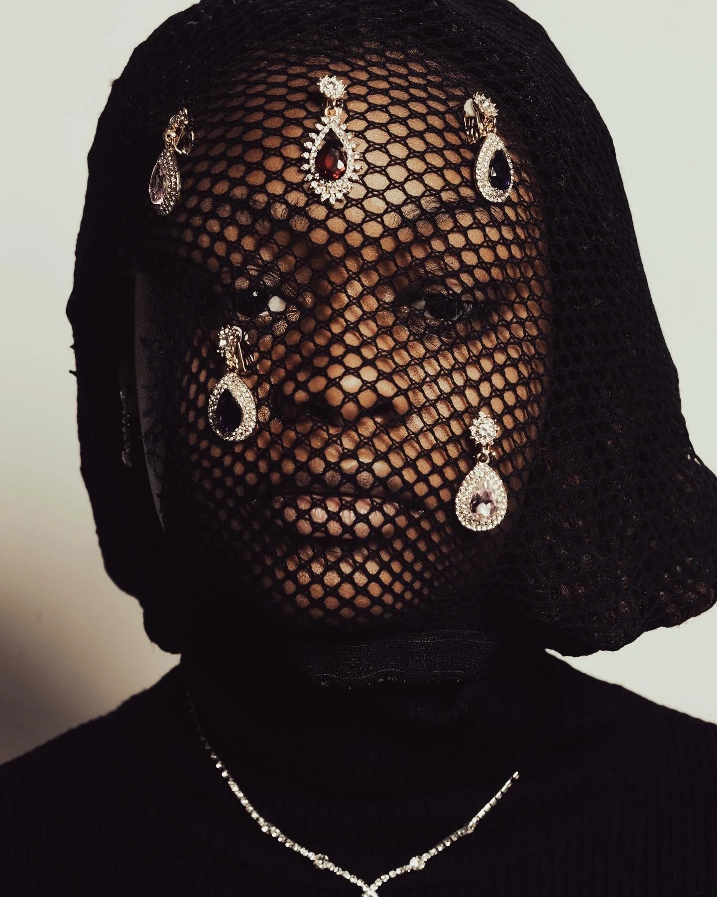 Person wearing black mesh head covering with jewelry, including earrings and a necklace, partially obscuring their face.