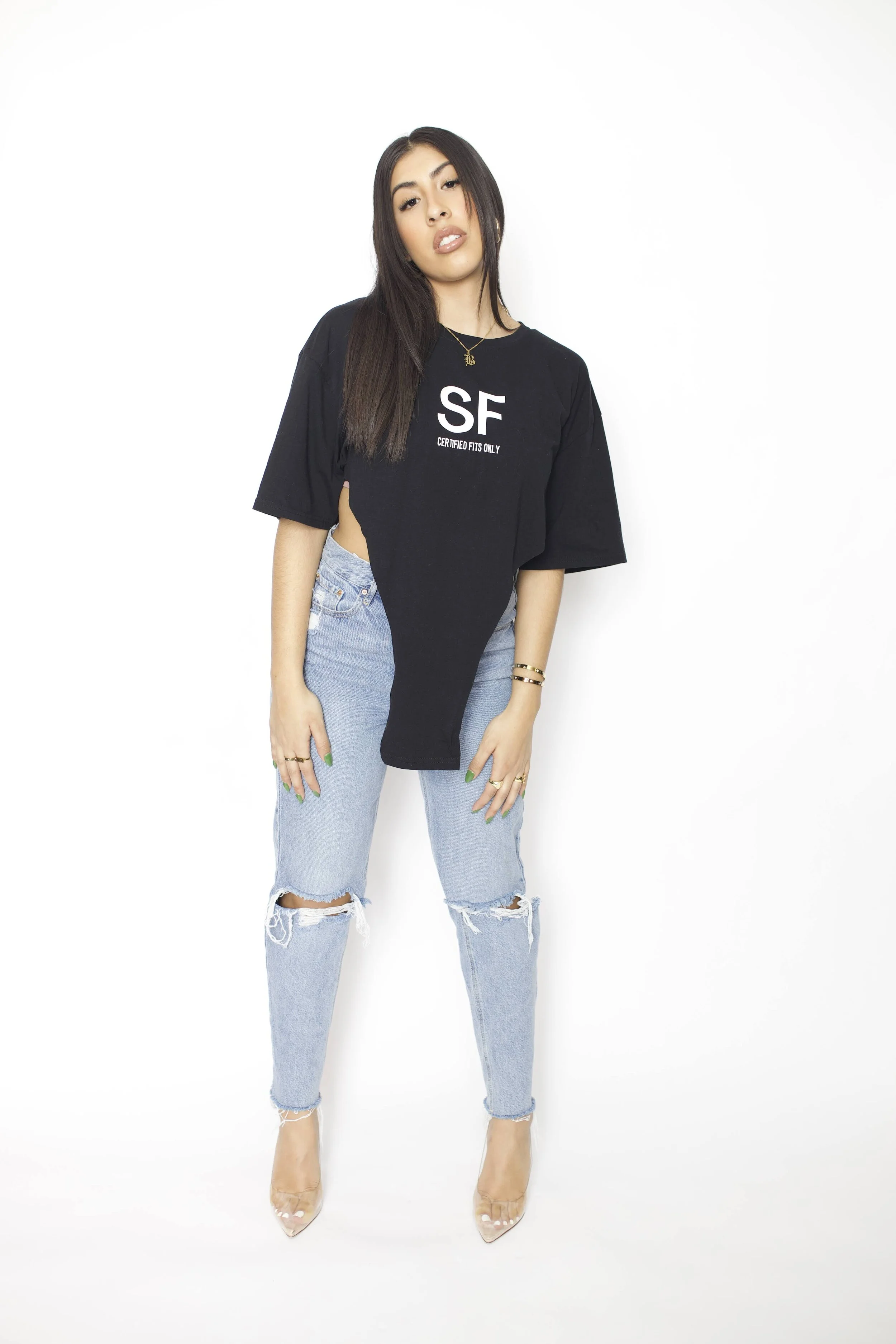 A woman with long dark hair wears a black oversized T-shirt, ripped blue jeans, and nude high heels, standing against a white background.