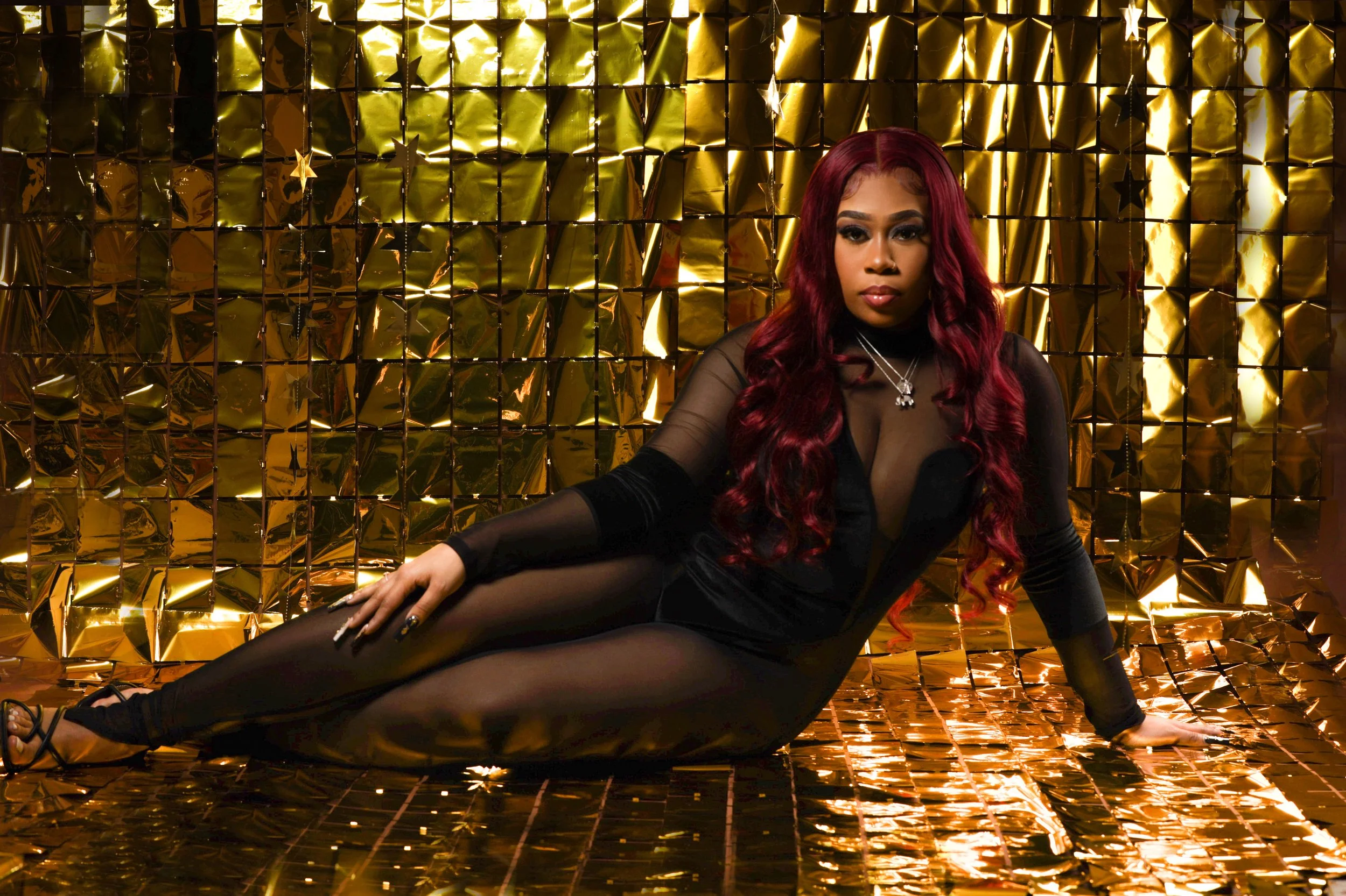 Woman with long red hair posing on a gold reflective backdrop in a black sheer dress.