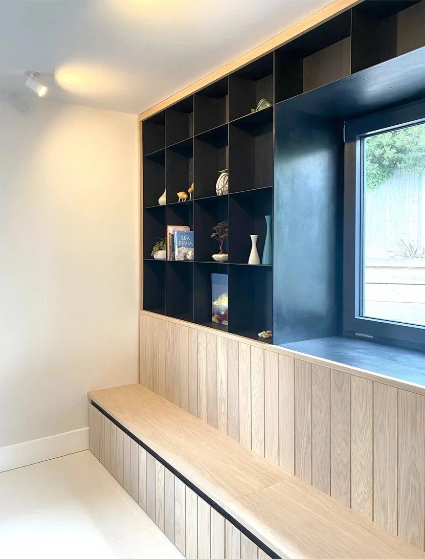 Photo of finished bespoke fitted oak bench seat joinery with shelving formed in black steel used to display colourful objects and wrapping around a window to frame views out