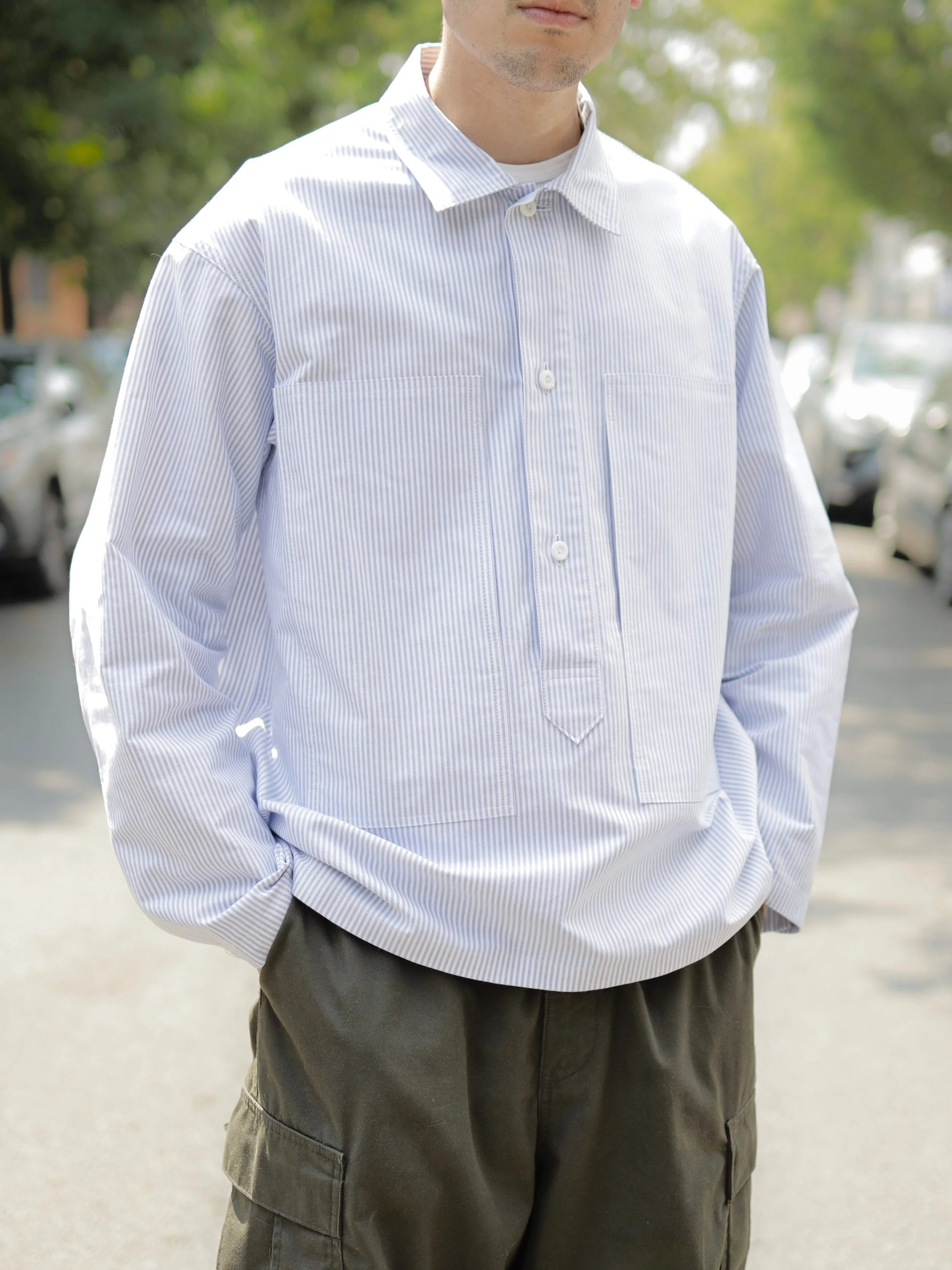 Blue striped Oxford cotton pullover shirt worn on body, featuring a relaxed fit, oversized pockets, and contemporary workwear-inspired styling.