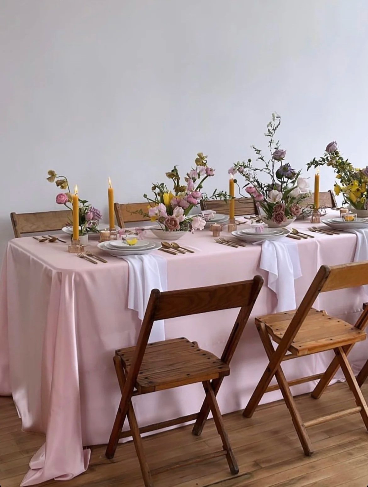 A dining table decorated with pink tablecloth, floral arrangements, white plates, gold utensils, pink flower napkins, and tall yellow candles; surrounded by wooden chairs.