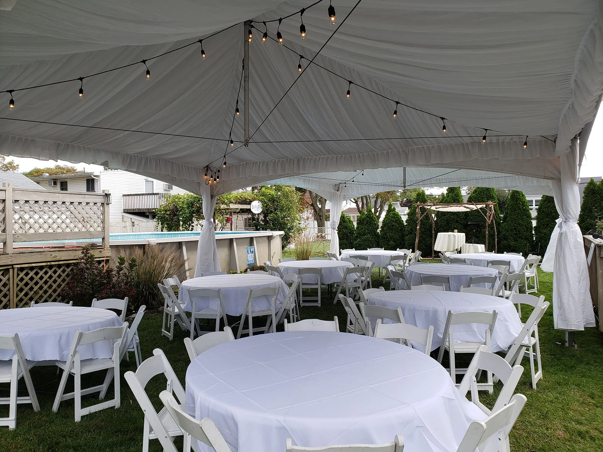 Outdoor event tent with round tables covered in white tablecloths and surrounded by white folding chairs. String lights hang from the tent ceiling, with trees and houses visible in the background.