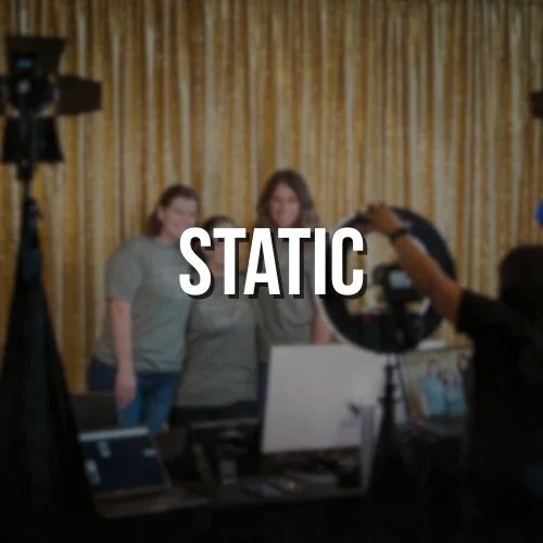 Two people posing for a photo in front of a gold curtain with a camera person taking their picture