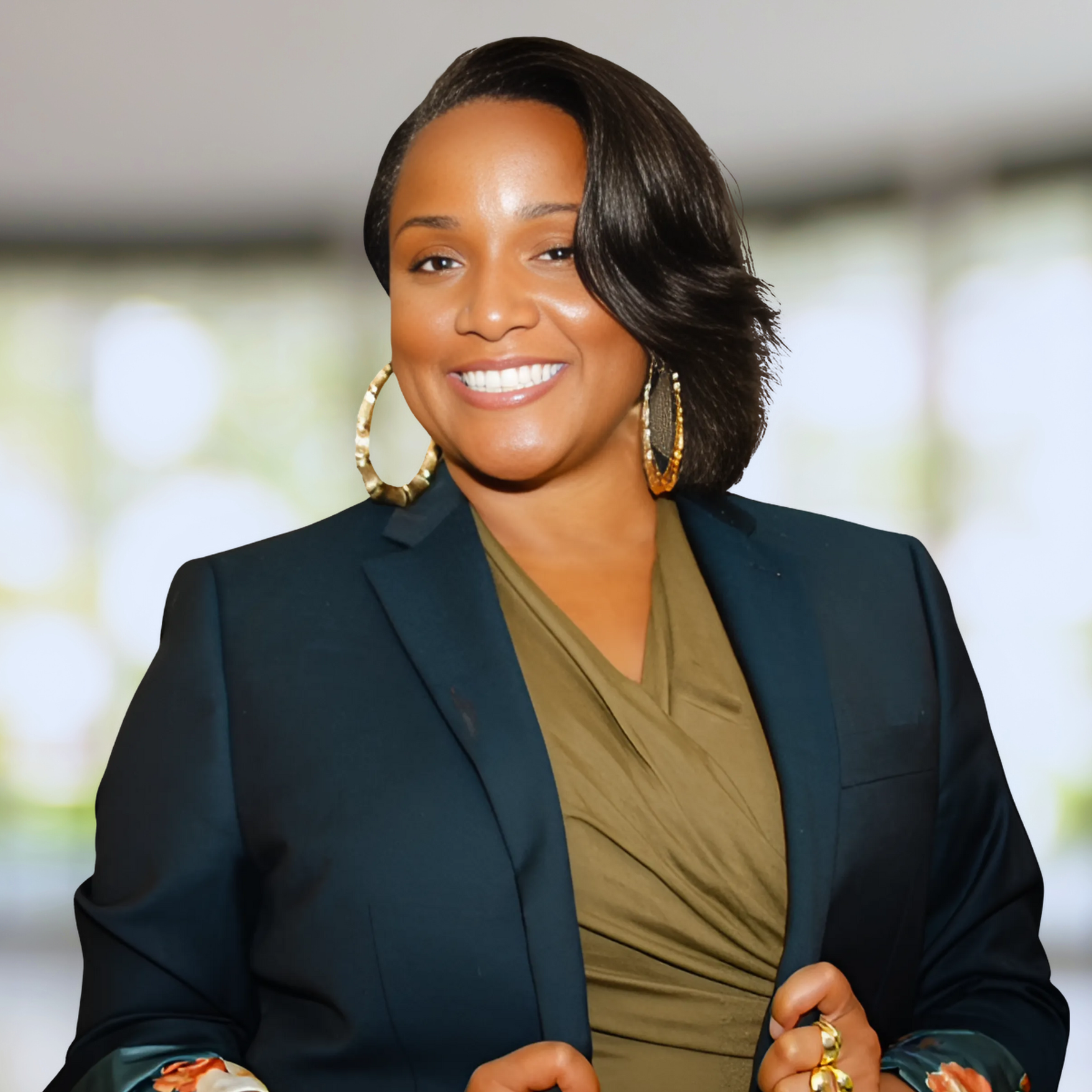 A smiling woman with shoulder-length dark hair, wearing a black blazer, olive green blouse, large gold hoop earrings, and rings, standing indoors with a blurred background.