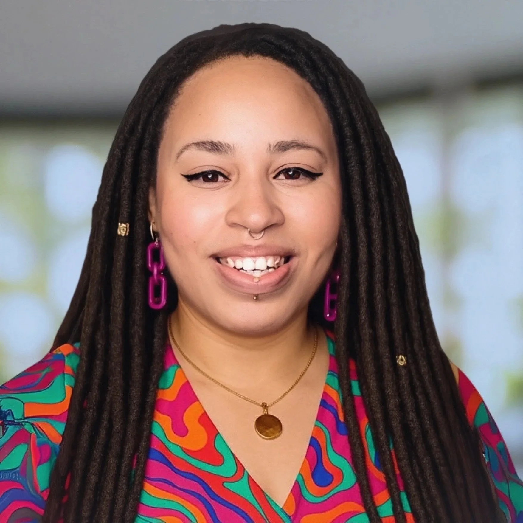 A young woman with long dreadlocks, multiple facial piercings, and bright makeup, smiling and looking at the camera, wearing a colorful patterned top, purple earrings, and a gold necklace, with a blurred indoor background.
