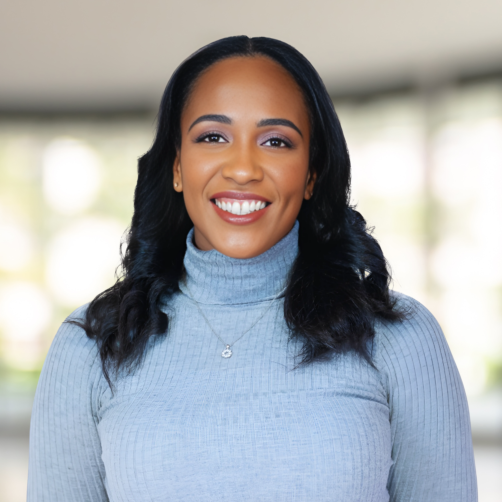 A woman with long black hair, wearing a light gray turtleneck sweater with a necklace, smiling at the camera with a blurred background of windows and greenery.