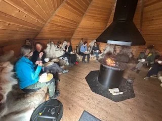 A snowy landscape with a line of sled dog teams and mushers, set against a backdrop of snow-covered mountains and a clear blue sky.
