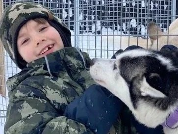 A smiling child in camouflage winter clothing and a hat is interacting with a husky puppy inside a fenced outdoor area during winter.