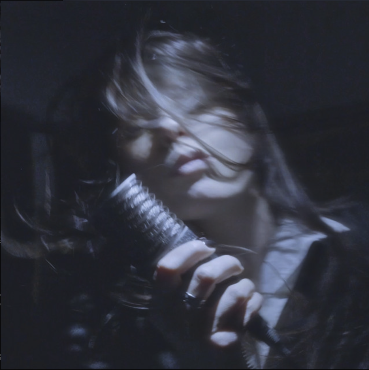 A woman with wind-blown hair, wearing leather gloves, holding a microphone, in a dark setting.