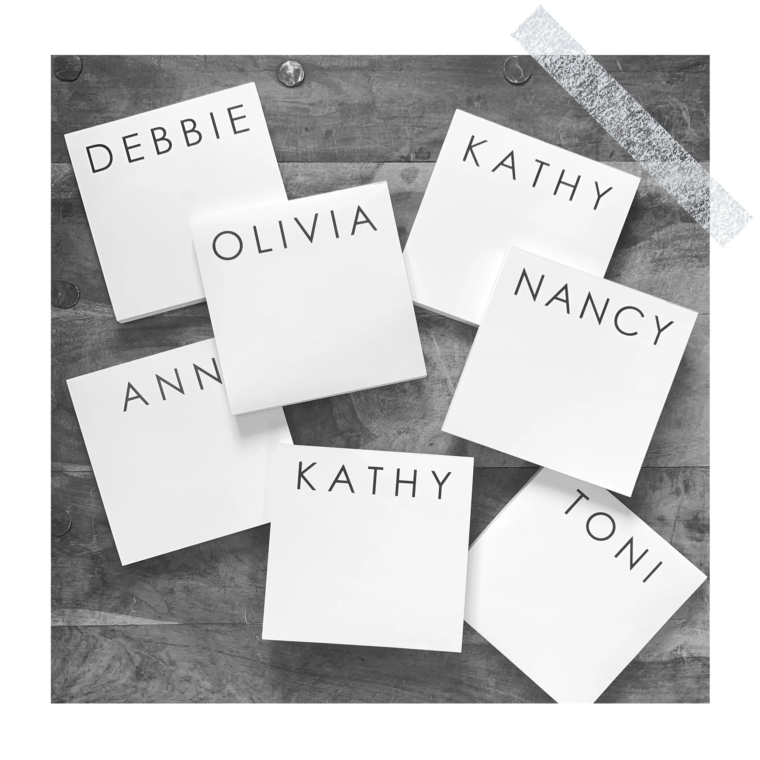 White sheets of paper with names written in bold uppercase letters, scattered on a wooden surface. Names include Debbie, Olivia, Kathy, Nancy, Ann, Toni.