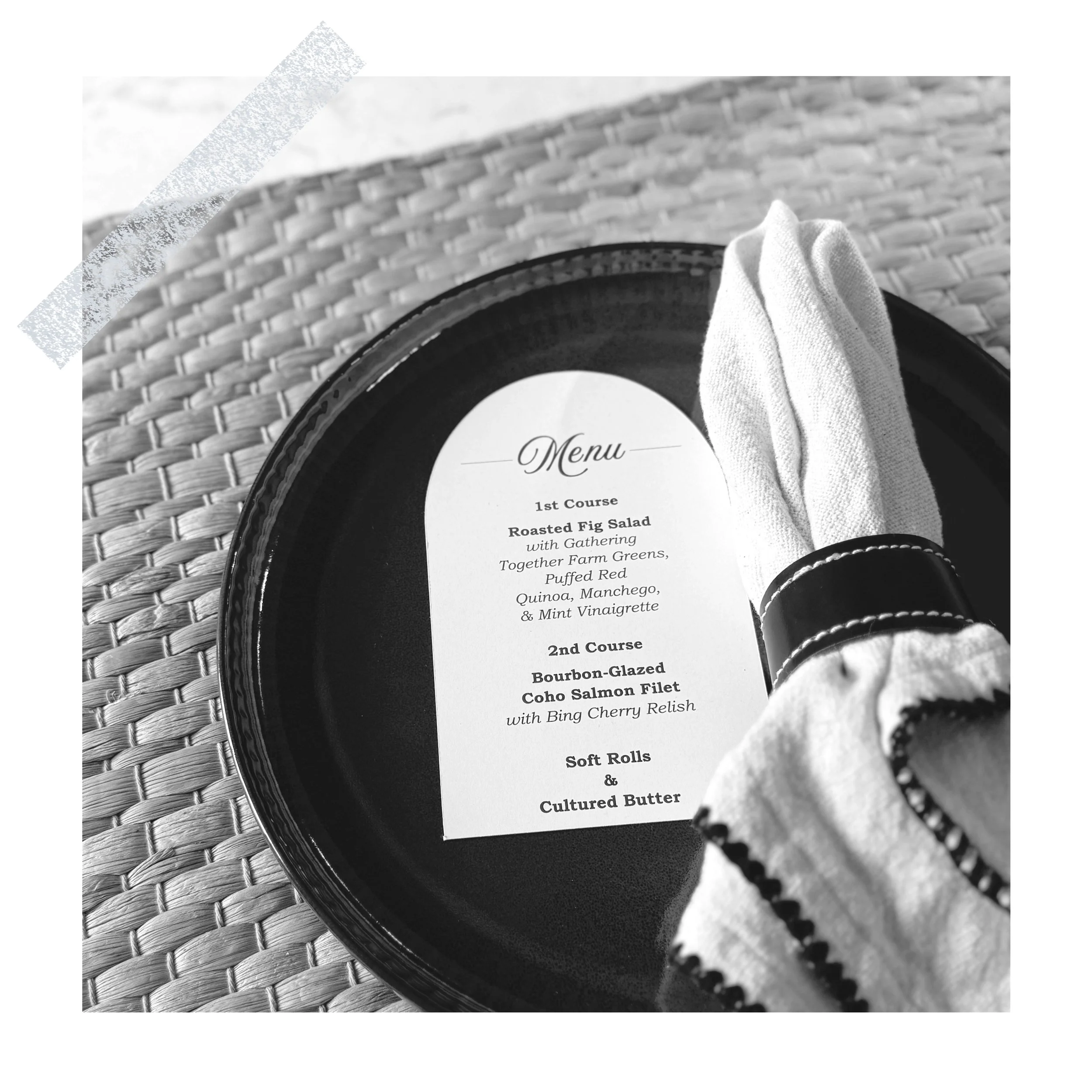 A black plate with a printed menu in the center, placed on a woven placemat, with a white cloth napkin wrapped with a black napkin ring on the side.