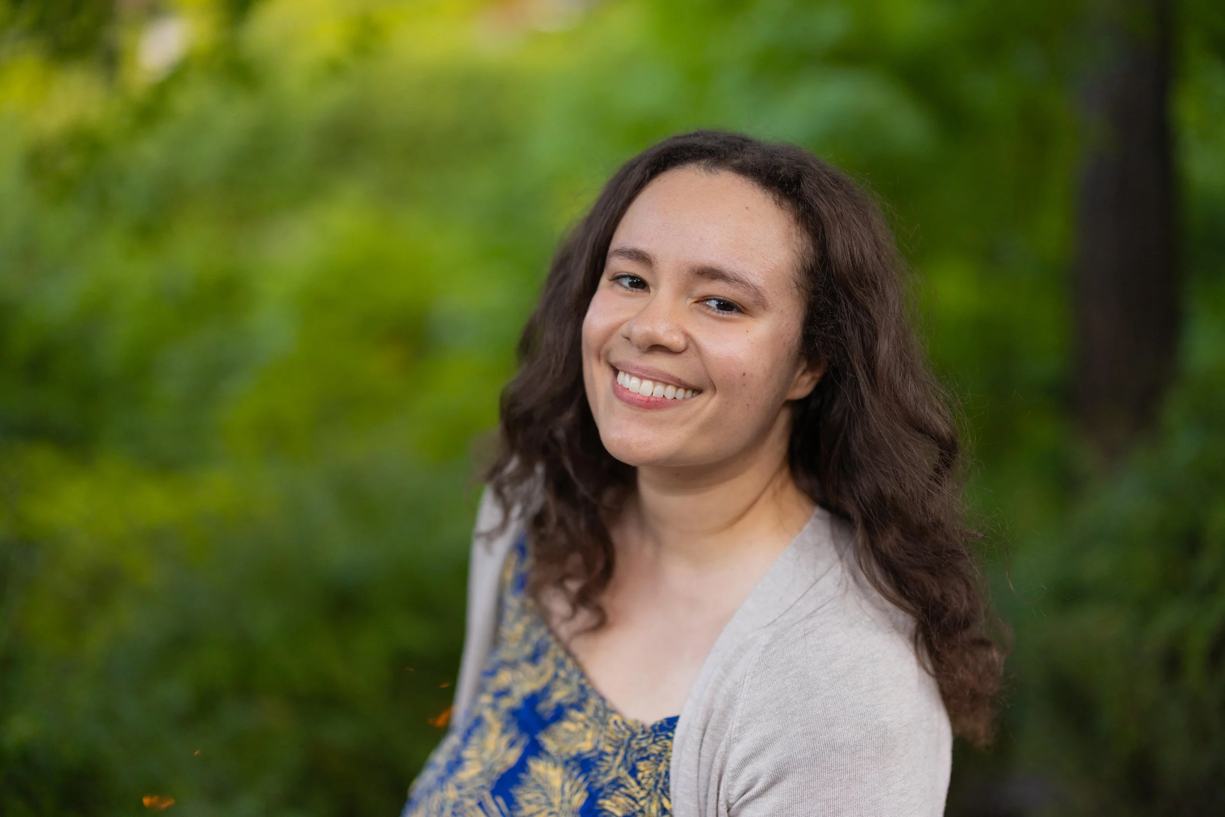 A young woman with long, wavy brown hair smiling outdoors with a blurred green background.