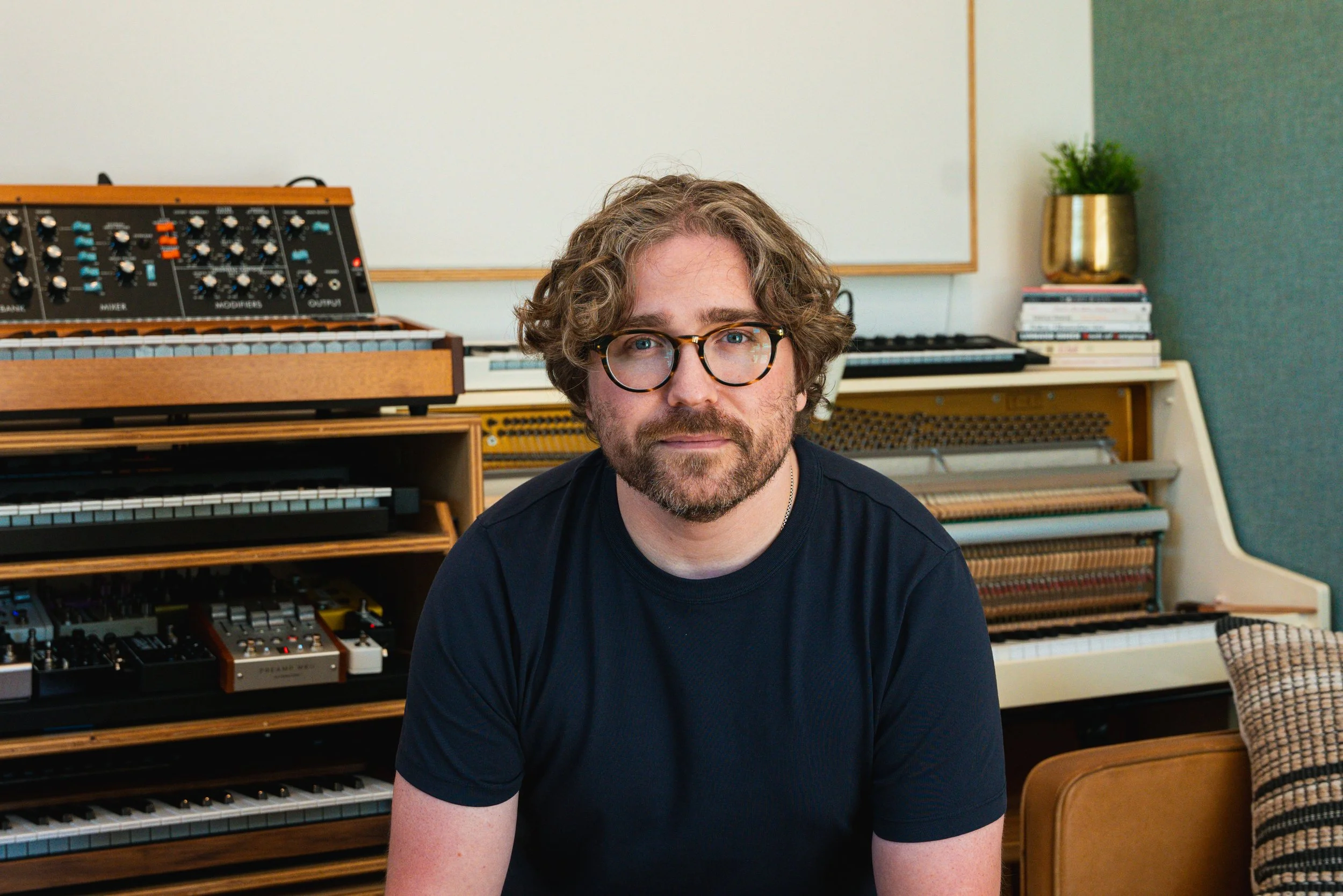A man with curly hair, glasses, and a beard sitting in front of a collection of musical instruments, including a keyboard, synthesizer, and various audio equipment, in a room with a whiteboard and a potted plant in the background.