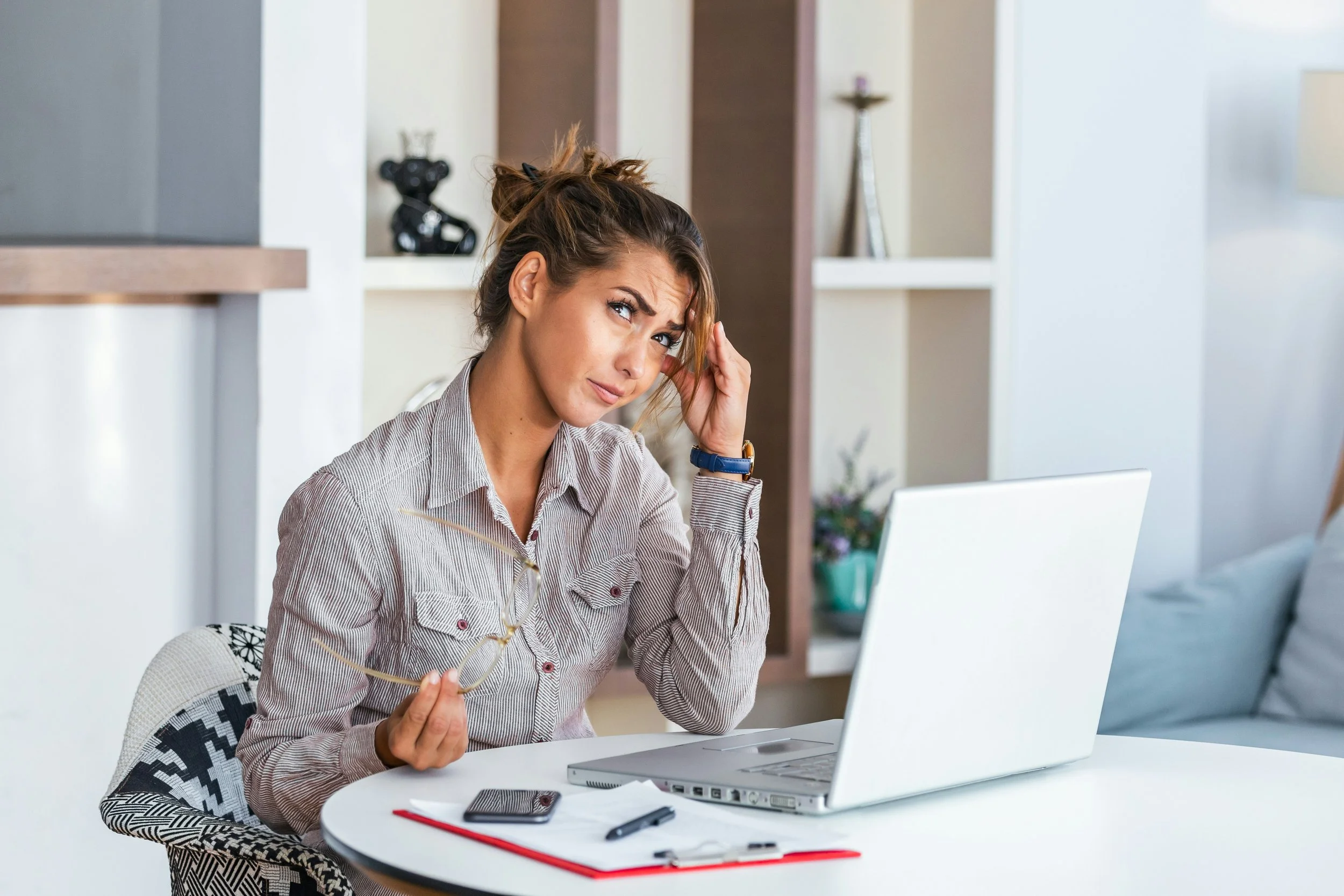 A woman sitting at a desk looking confused while holding glasses and touching her head, with a laptop and notepad in front of her in a bright room.