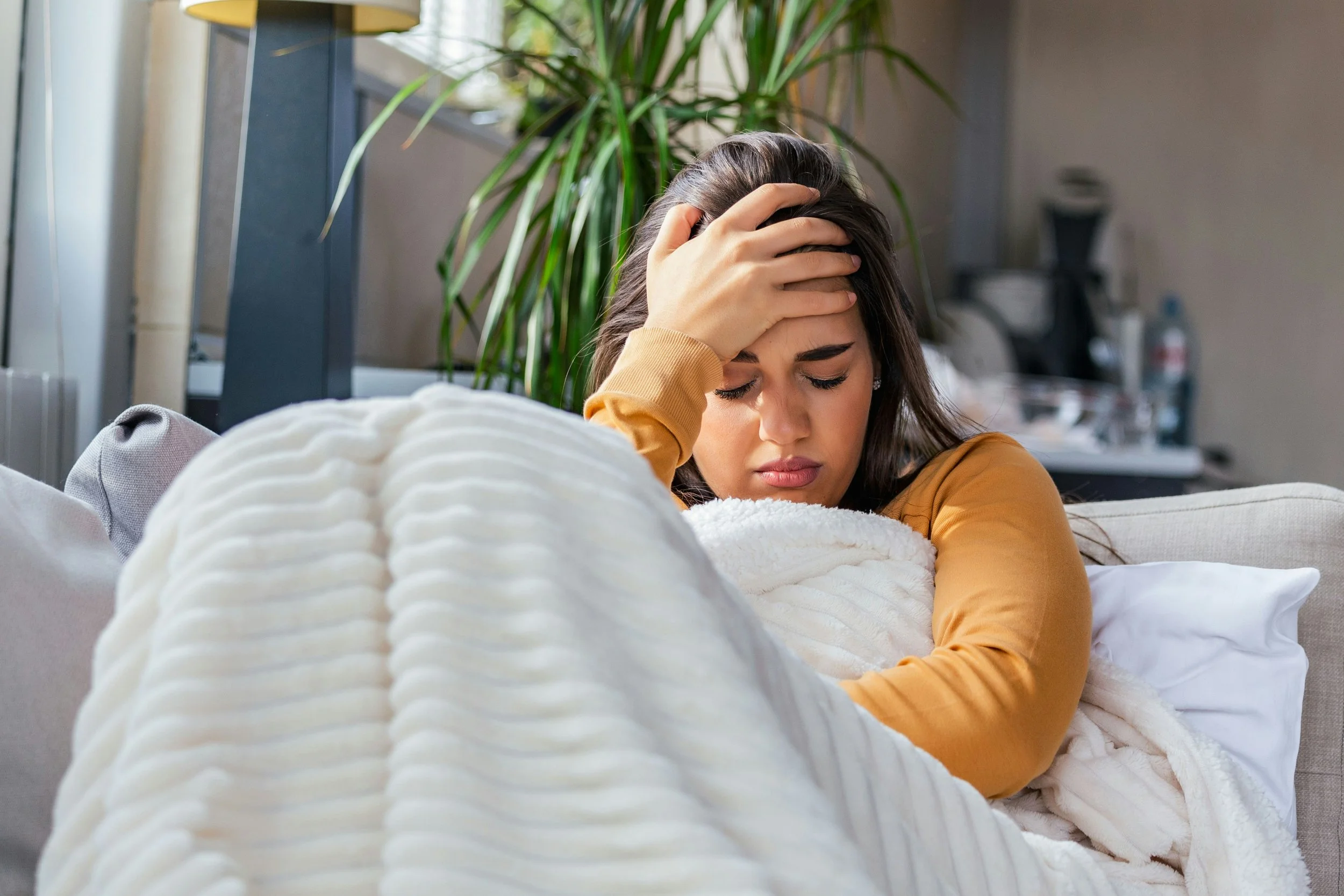 A woman sitting on a sofa, holding her head with a worried or distressed expression, with a blanket wrapped around her, in a room with a large plant and kitchen appliances in the background.