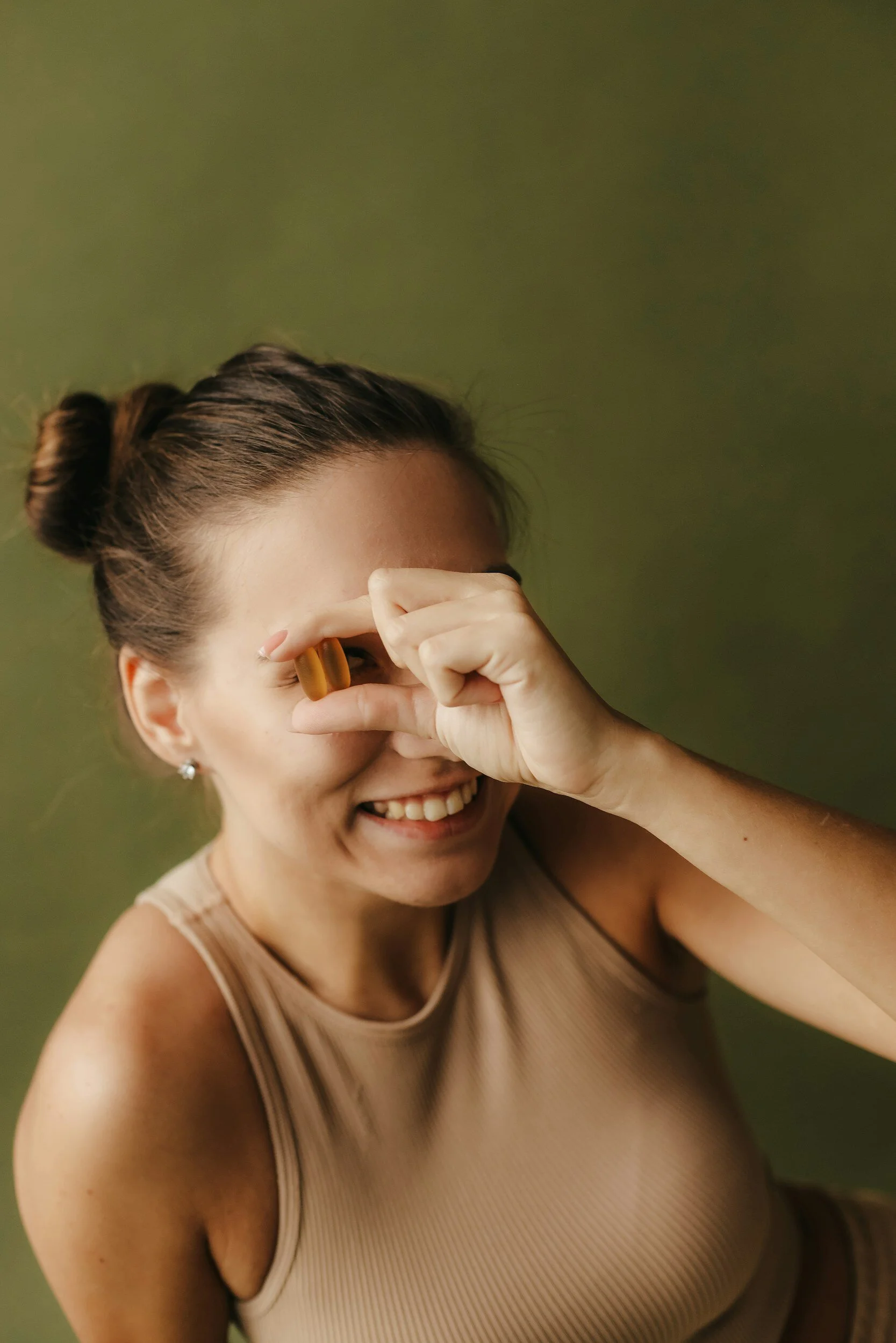 A woman smiling and making a frame with her fingers around her eye, wearing a beige sleeveless top and earrings, with brown hair pulled back in a bun, against a green background.