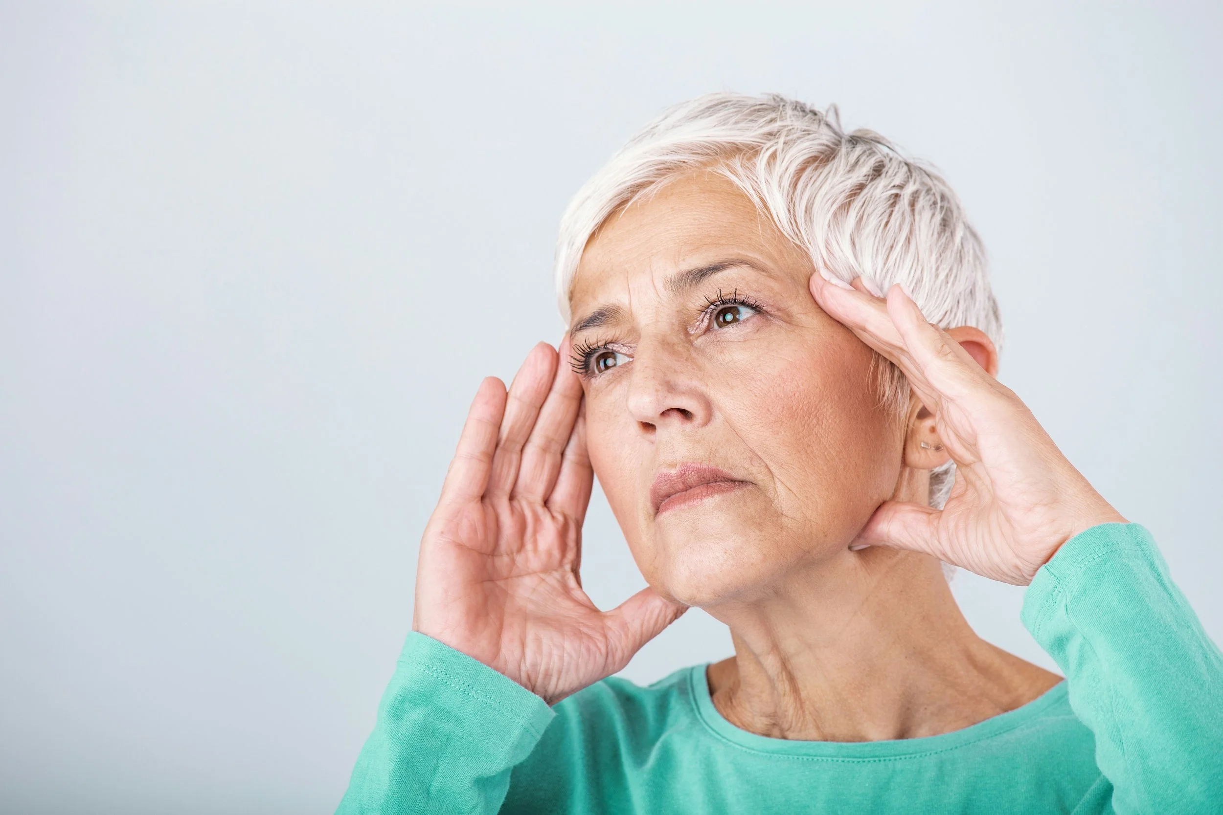 An elderly woman with short white hair holding her temples and looking upward with a thoughtful or distressed expression.