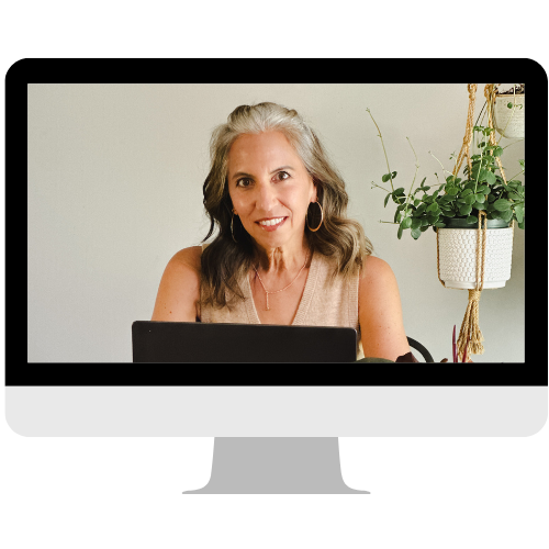 A smiling woman with gray hair sitting at a desk in front of a laptop, with a plant hanging on the wall behind her.