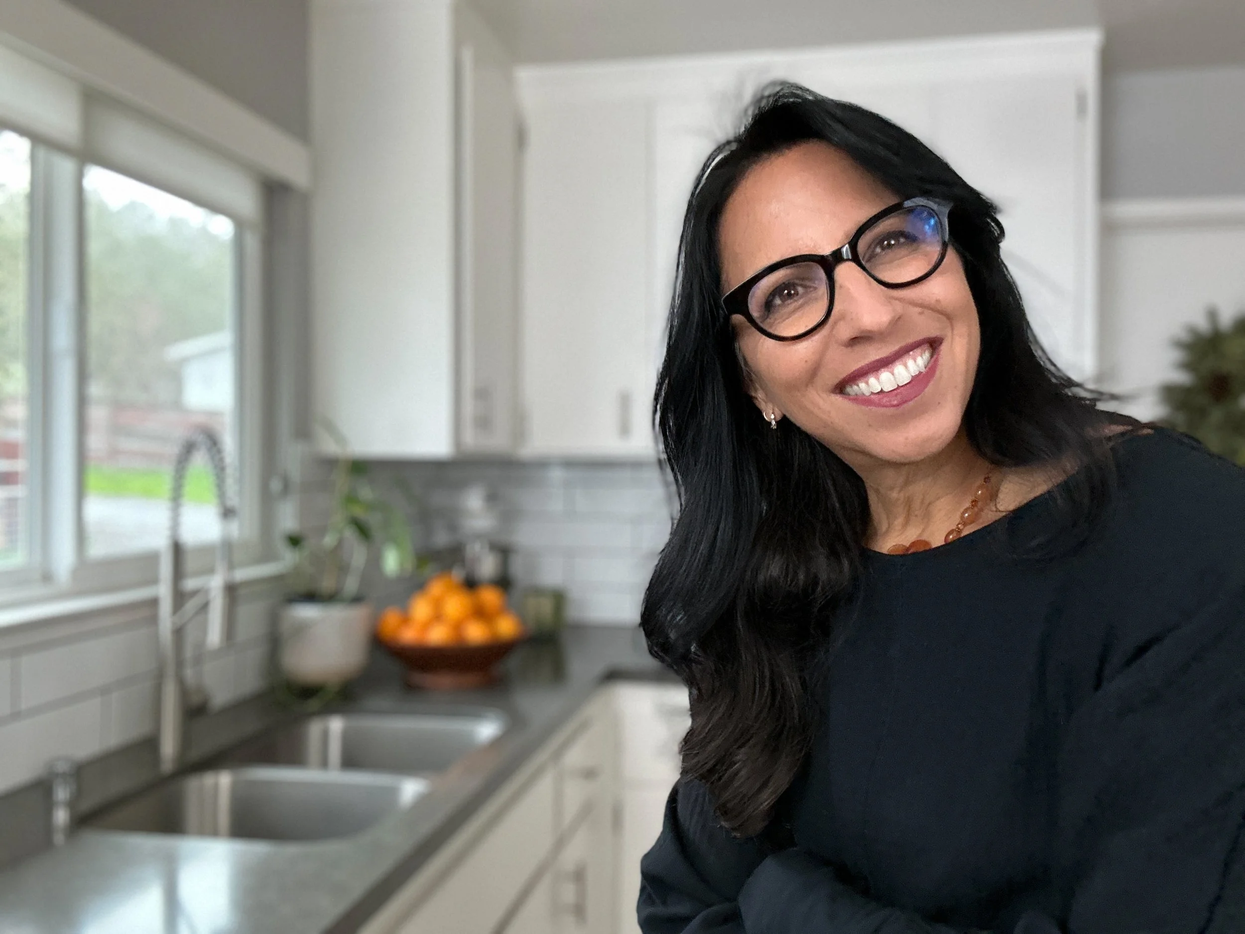 A woman with long dark hair and glasses smiling in a kitchen with white cabinets and window, with a bowl of oranges on the counter.