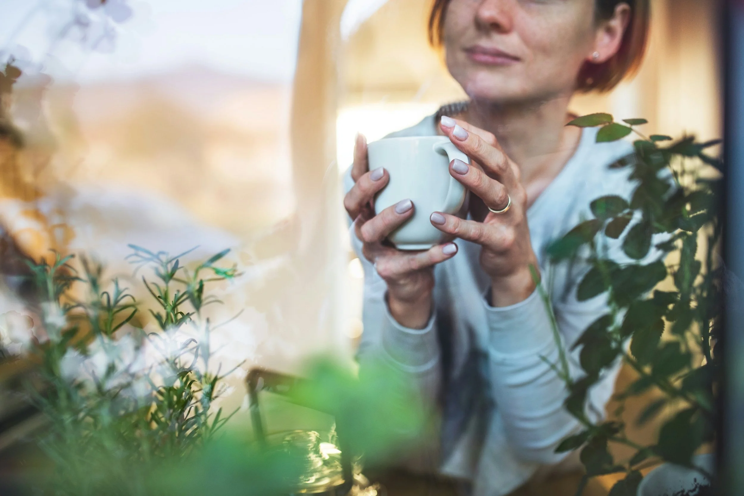 Woman holding a white mug with both hands, looking out a window with green plants in foreground and blurred background