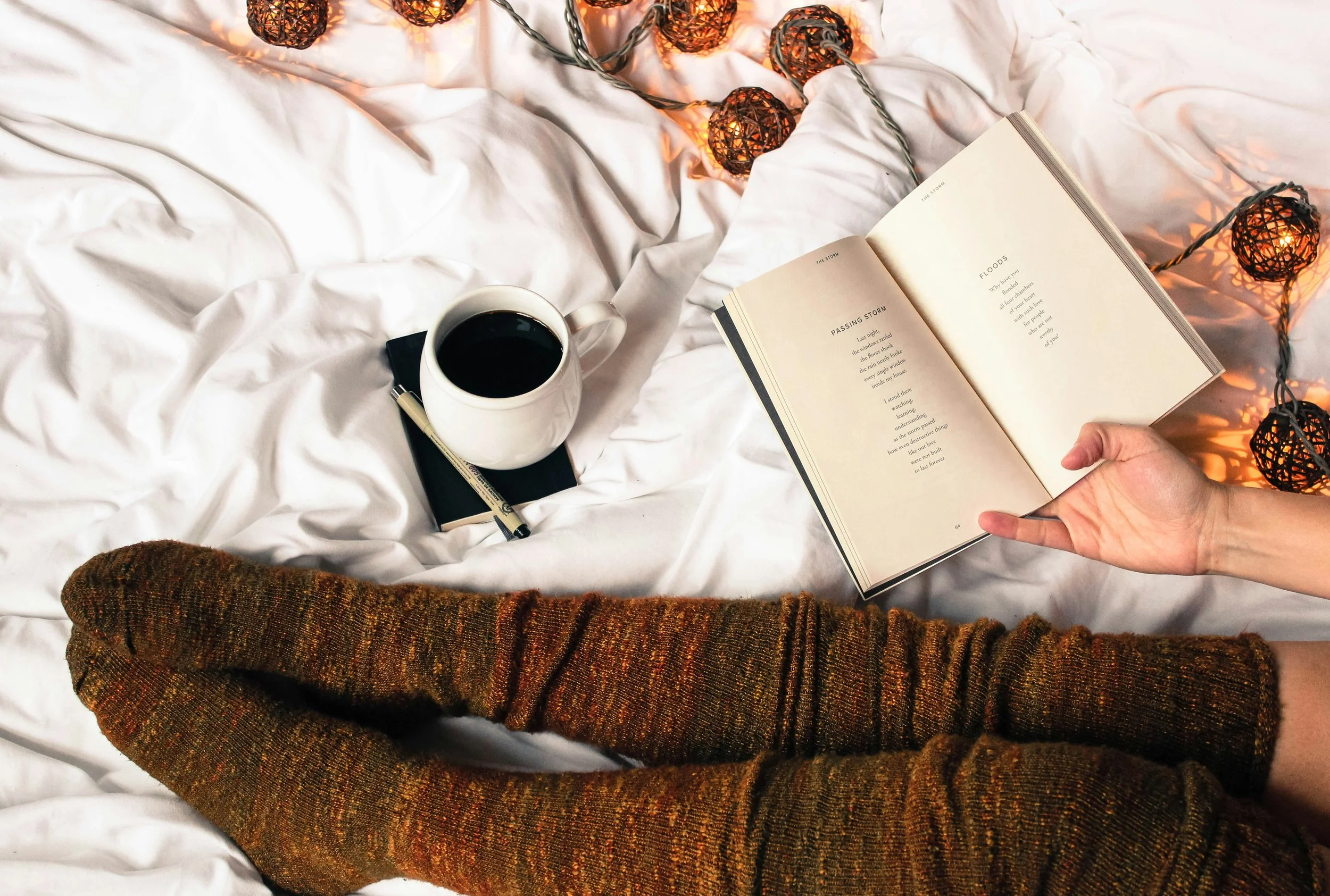 A person wearing warm, orange-brown knitted socks sits on a white, wrinkled bedspread. They hold an open book with poetry. Nearby, there is a cup of black coffee, a black notebook, and a pen on the bed. A string of decorative warm-colored lights is draped across the bed, creating a cozy ambiance.