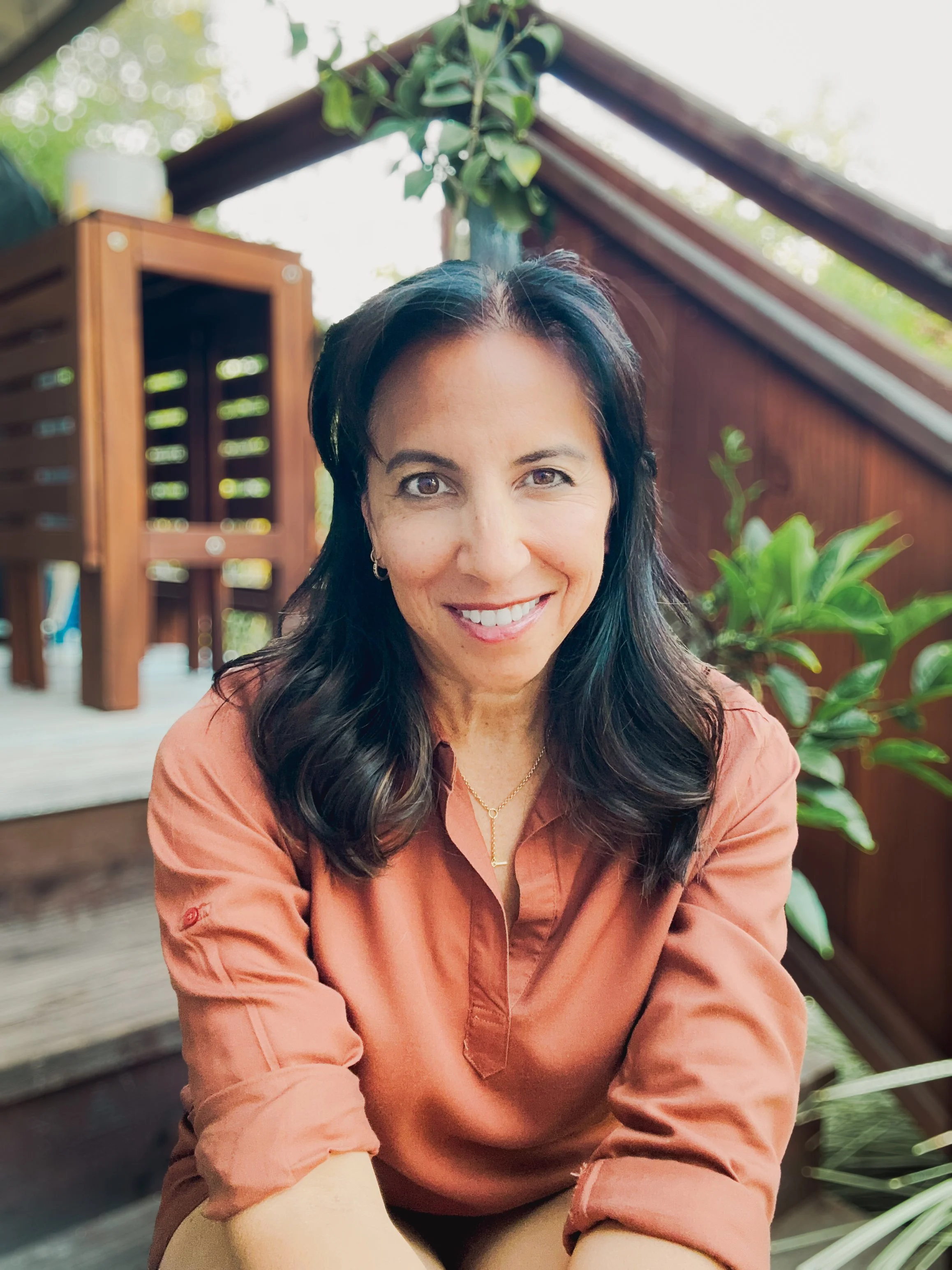 Smiling woman with dark hair wearing a rust-colored blouse, sitting outdoors in front of a wooden fence and plants.