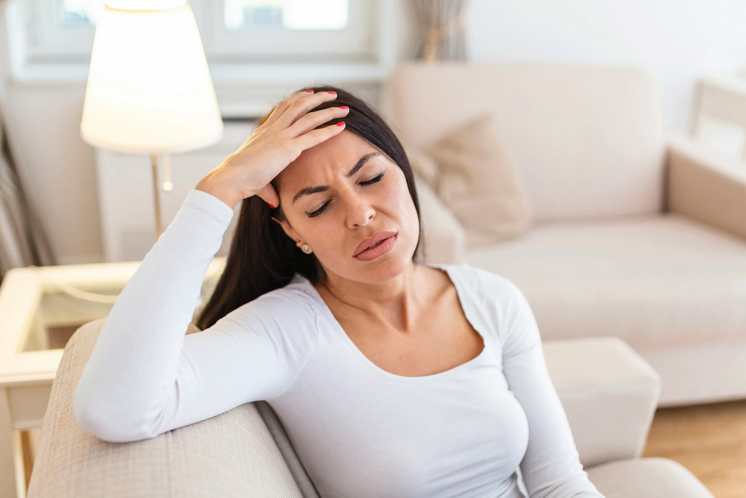A woman sitting on a beige couch with her eyes closed, holding her forehead with one hand, appearing to have a headache or feeling unwell in a bright living room