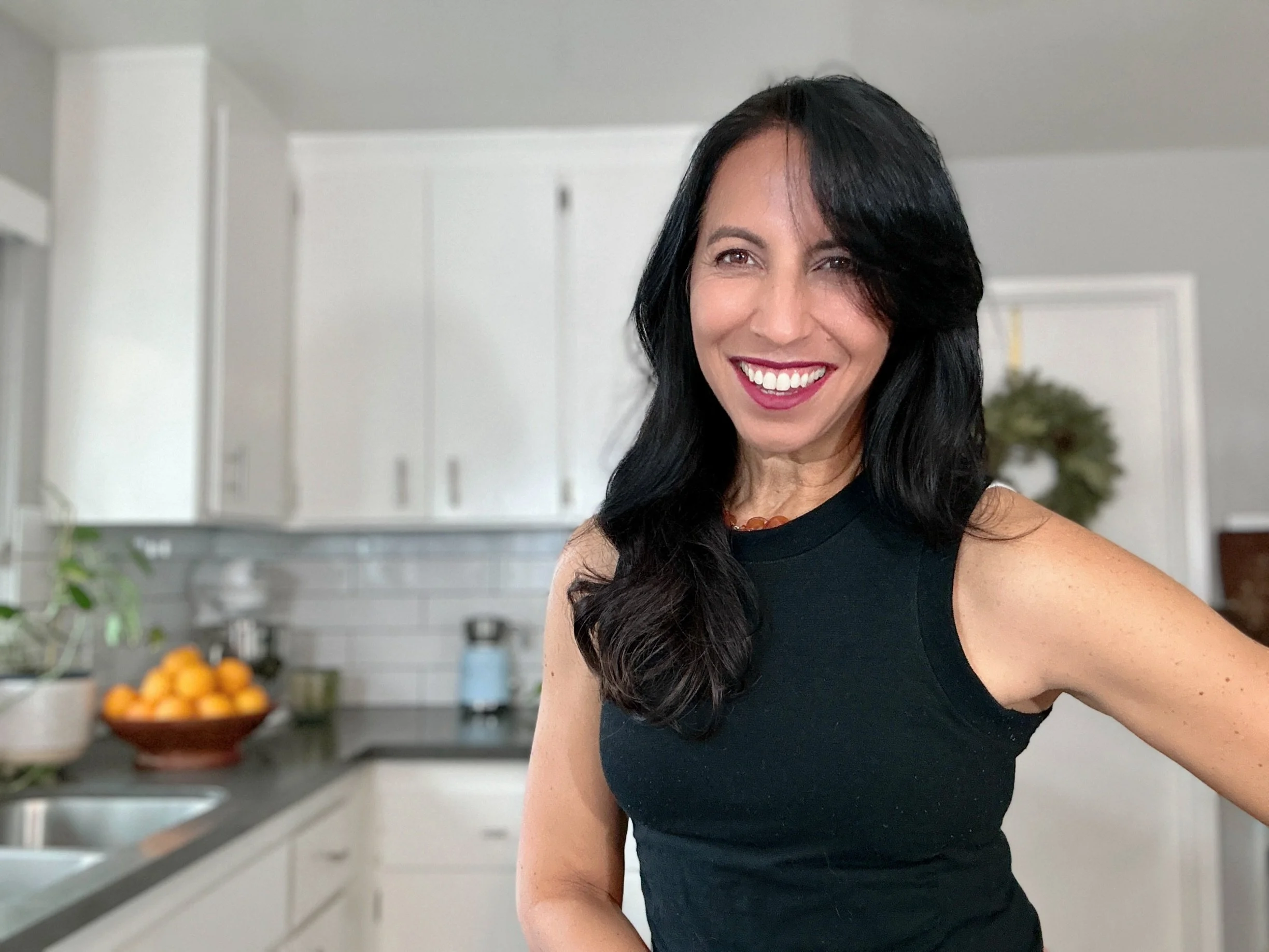 A woman with long black hair smiling and wearing a black sleeveless top in a modern kitchen.