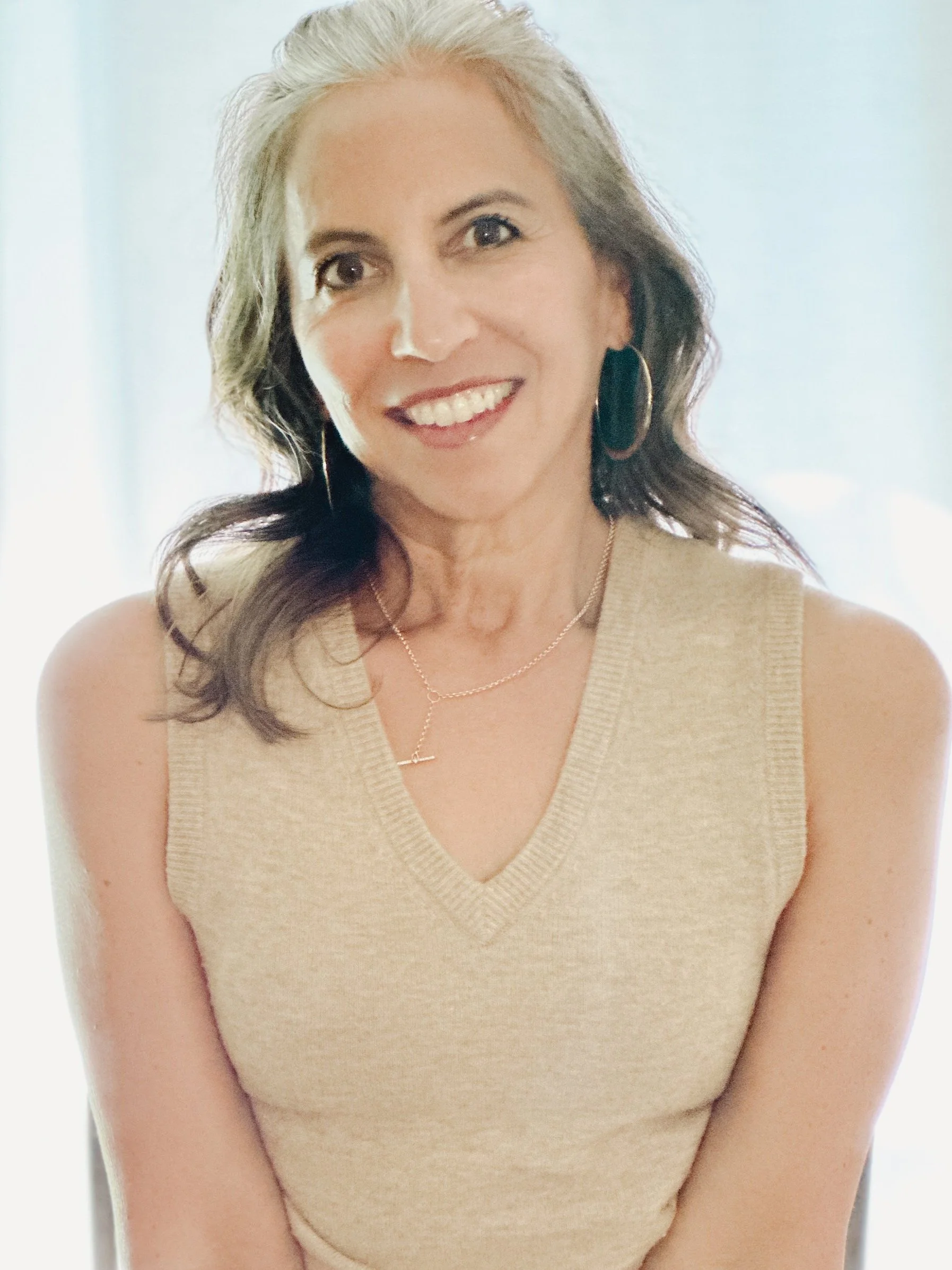 A woman with shoulder-length wavy brown hair and gray streaks, wearing a beige sleeveless V-neck top, hoop earrings, a delicate necklace, and smiling at the camera.