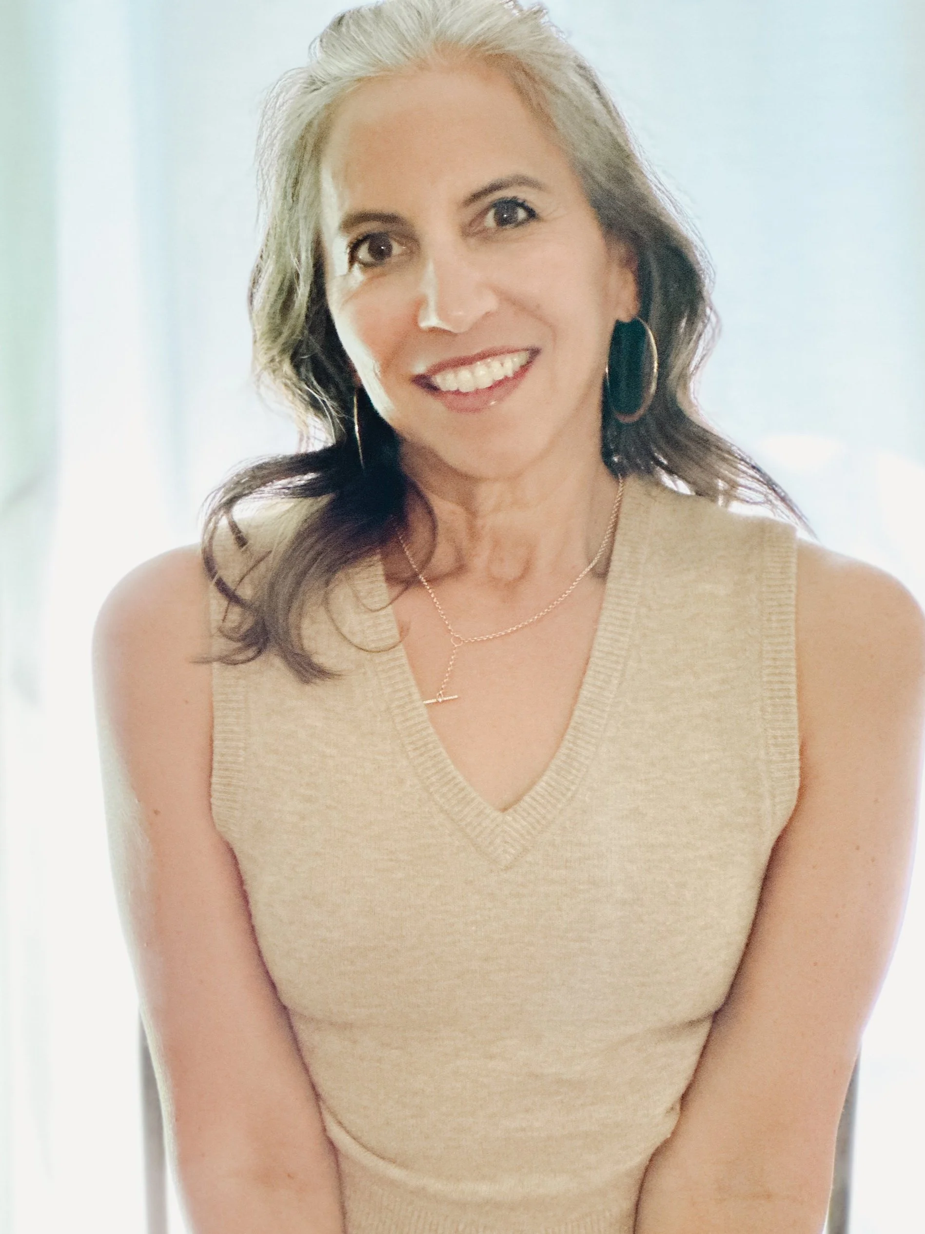 A woman with shoulder-length wavy brunette hair and light skin, wearing a beige sleeveless V-neck sweater, gold hoop earrings, a delicate gold necklace, and smiling at the camera.