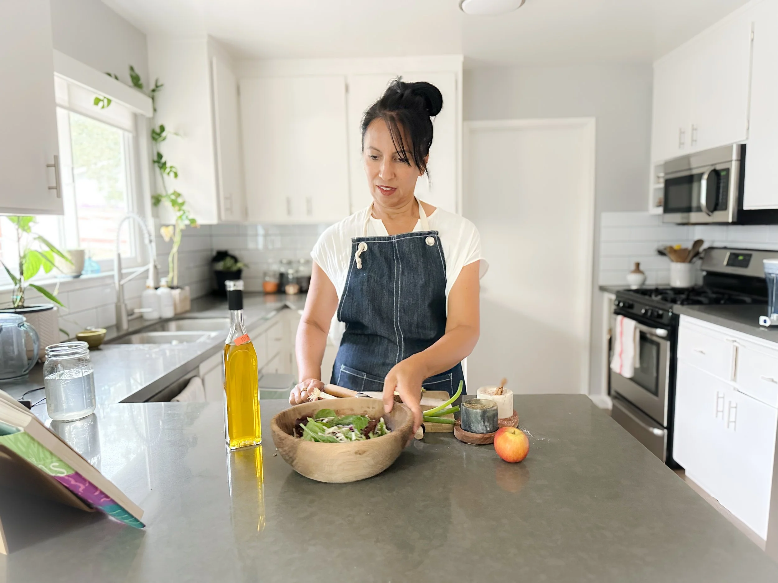 A woman in a white shirt and denim apron preparing a salad in a kitchen with white cabinets, a black stove, and a window with greenery outside.