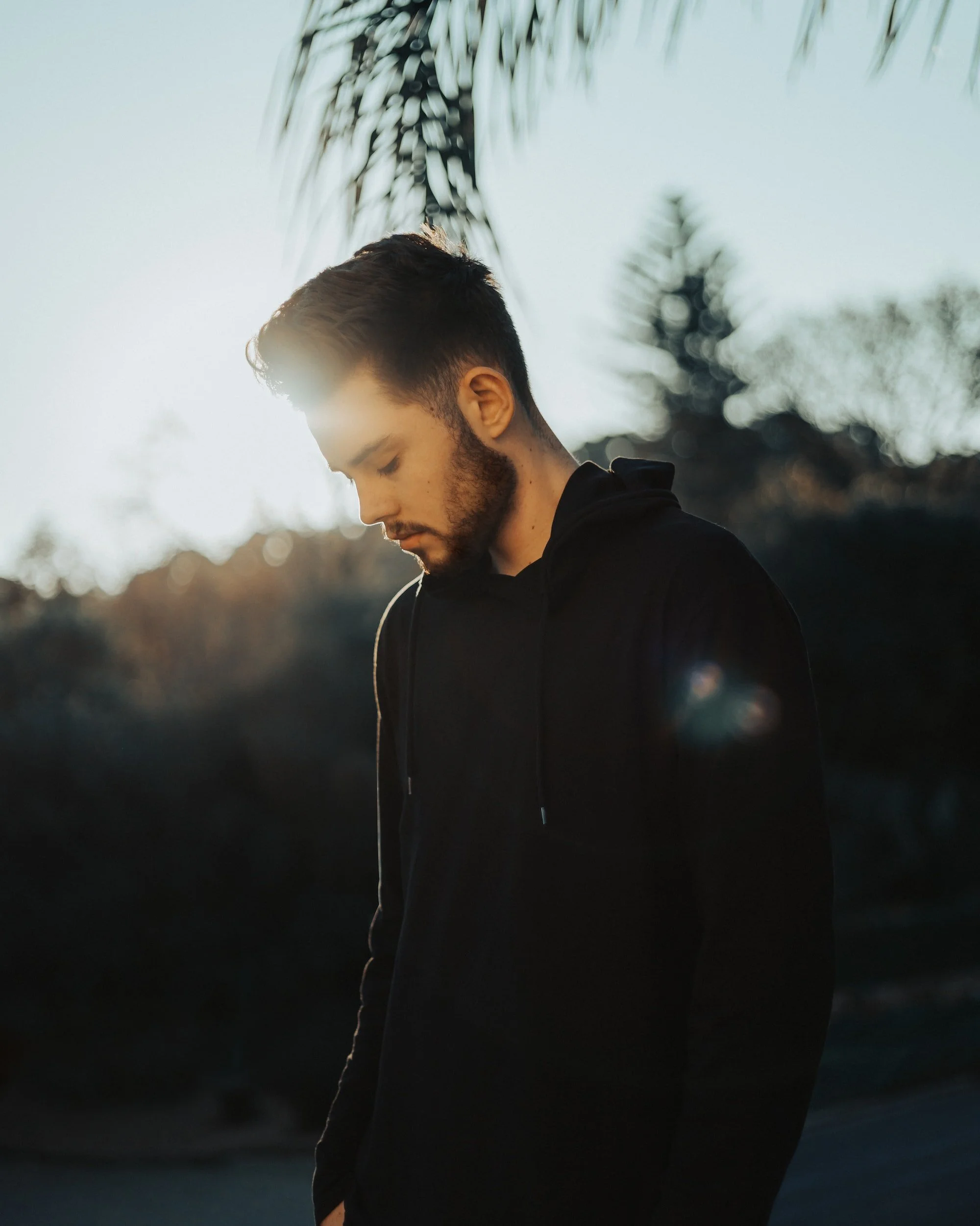 A young man with a beard in a black hoodie standing outdoors at sunset, looking down with a sun flare in the background.