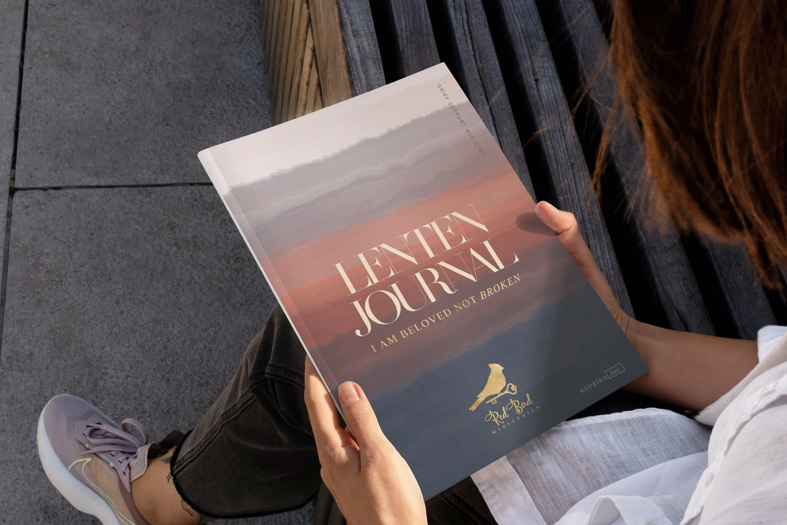 Person sitting on a bench reading a publication titled 'Lenten Journal, I Am Beloved Not Broken' with a sunset or sunrise scene on the cover.